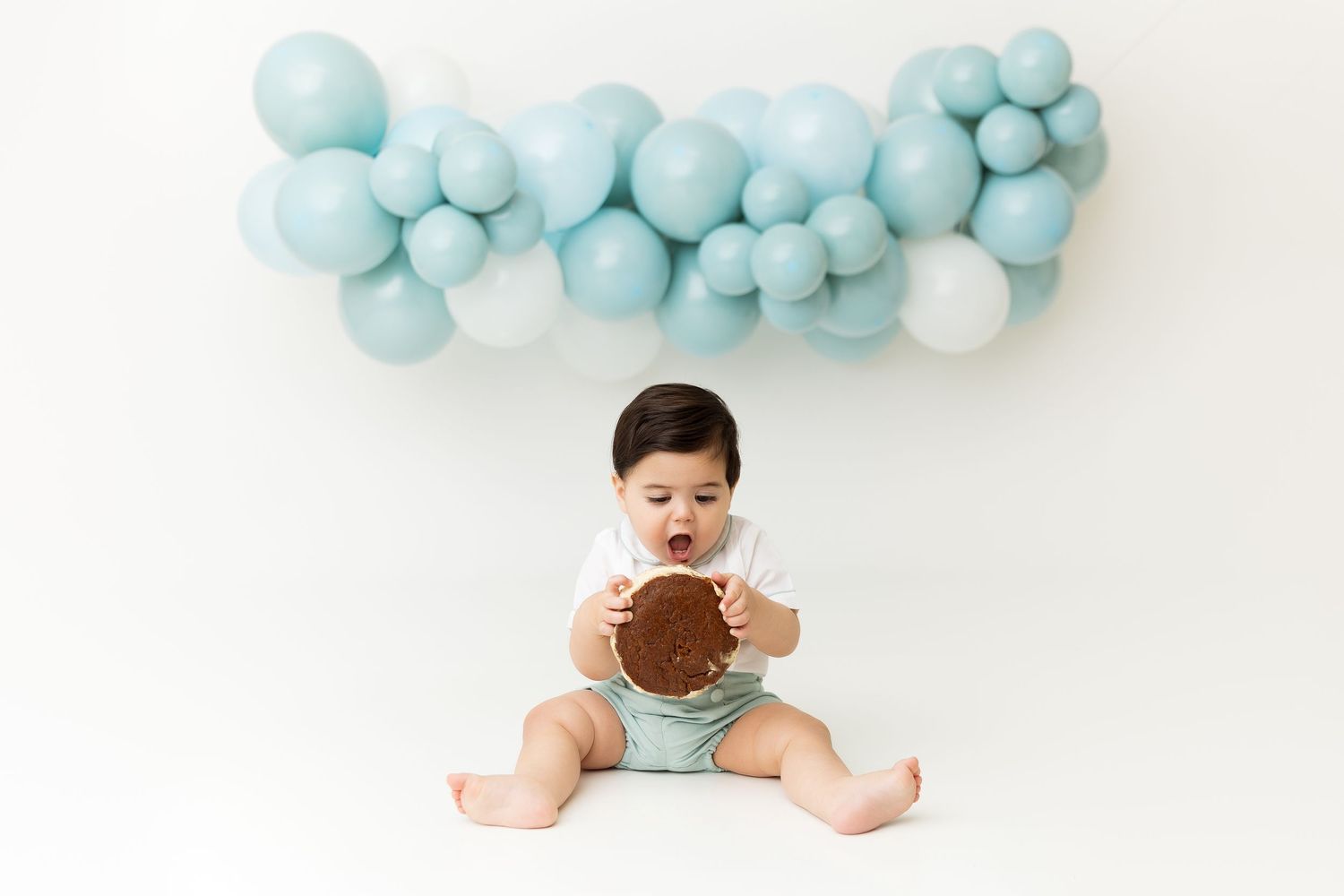 Baby milestone photo with blue balloon arch and chocolate cookie prop against white background.