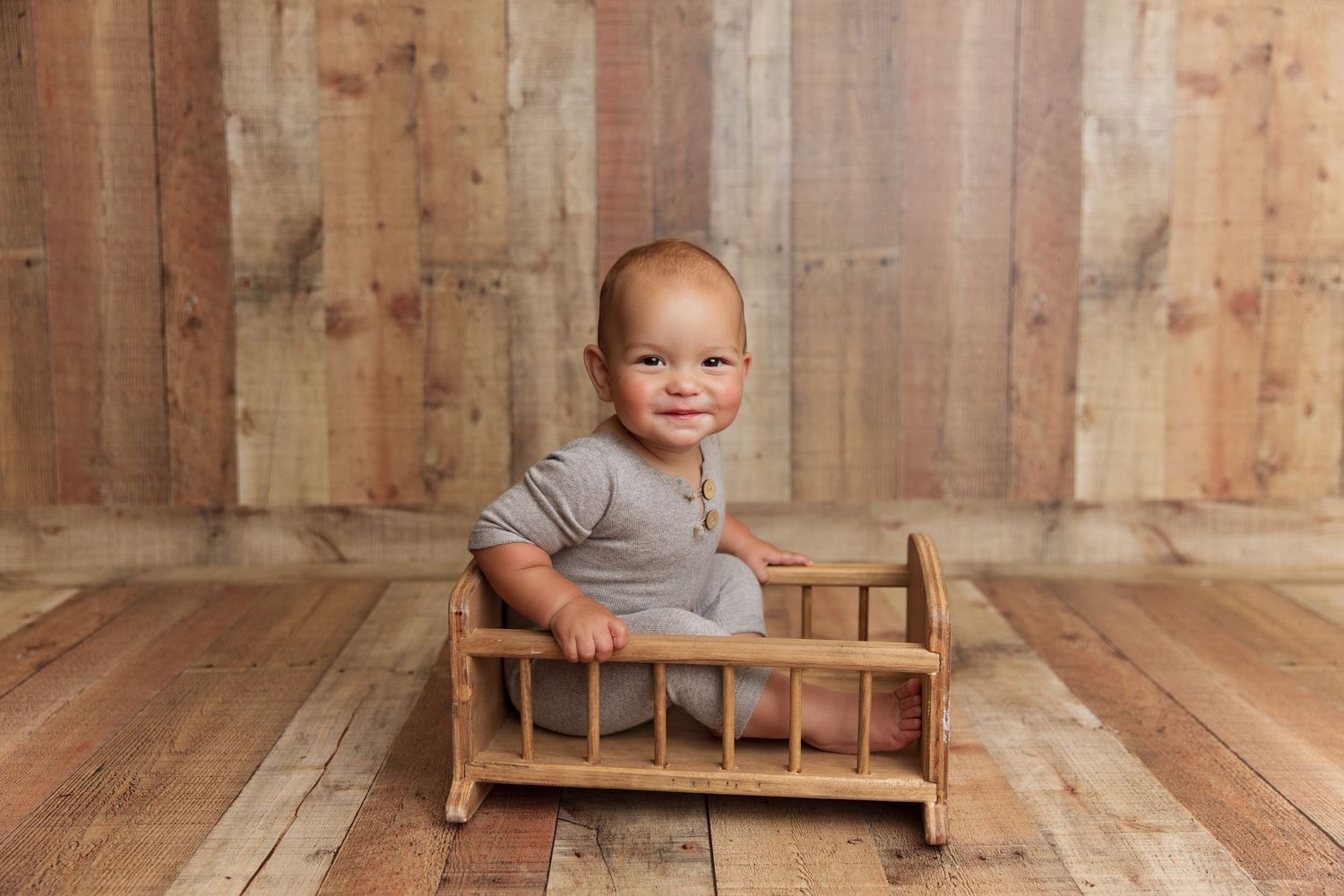 Baby in grey outfit sits happily in small wooden cradle on hardwood floor.