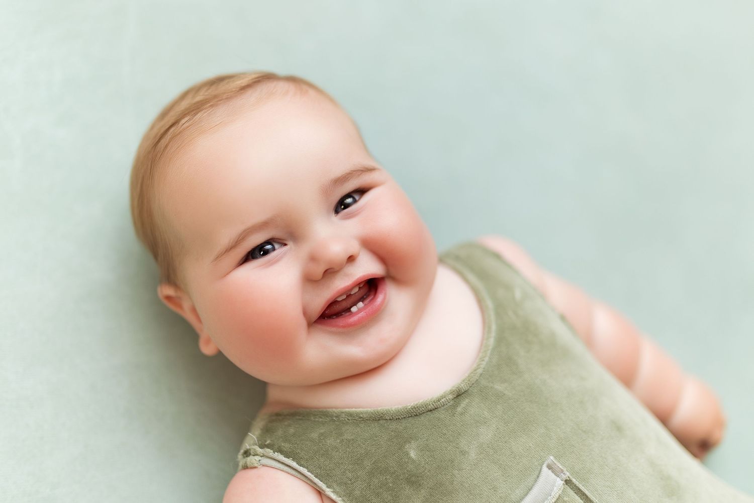 Happy baby in green outfit smiles against soft mint green background.