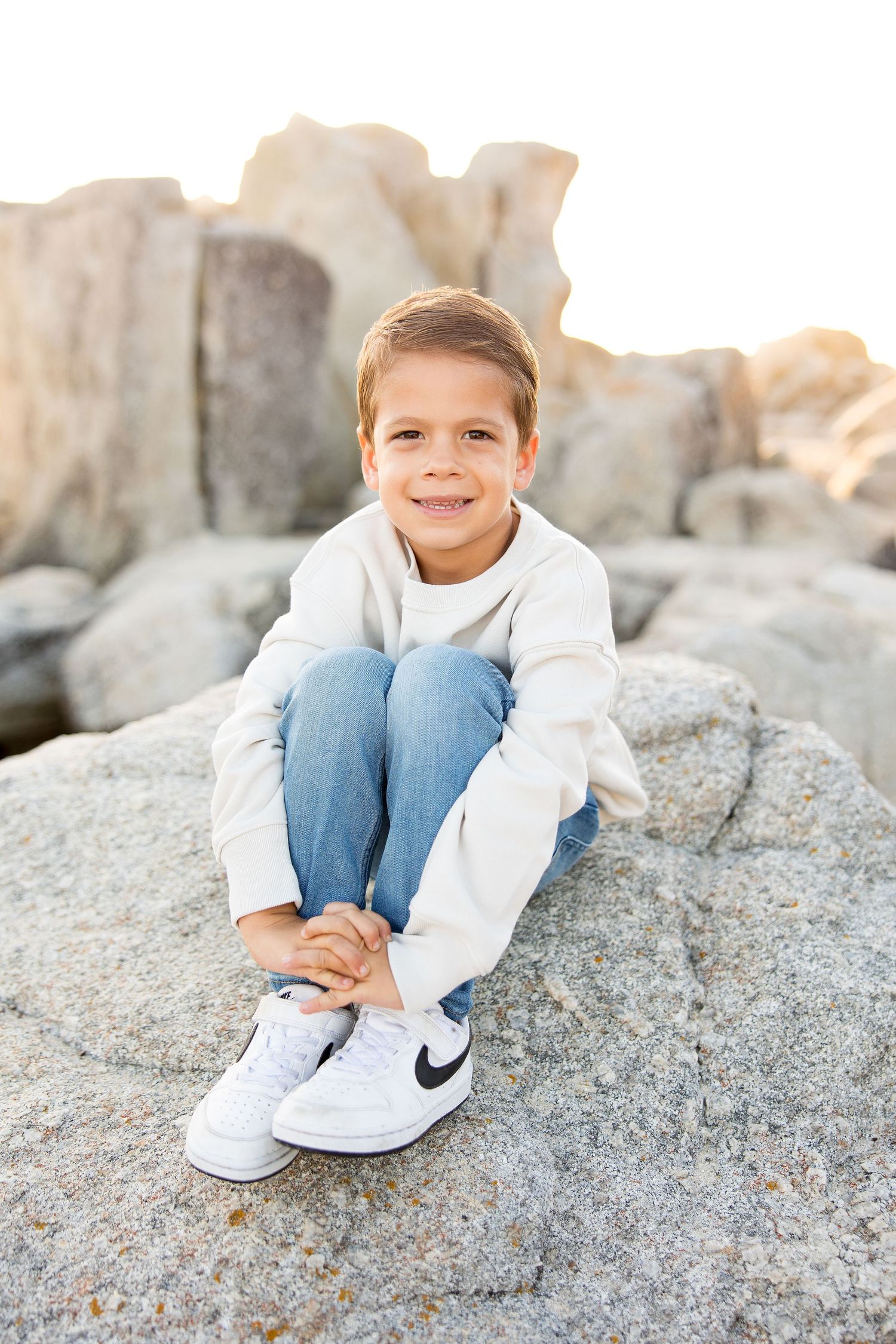 Young child in white sweater and Nike shoes sitting on coastal rocks during golden hour.