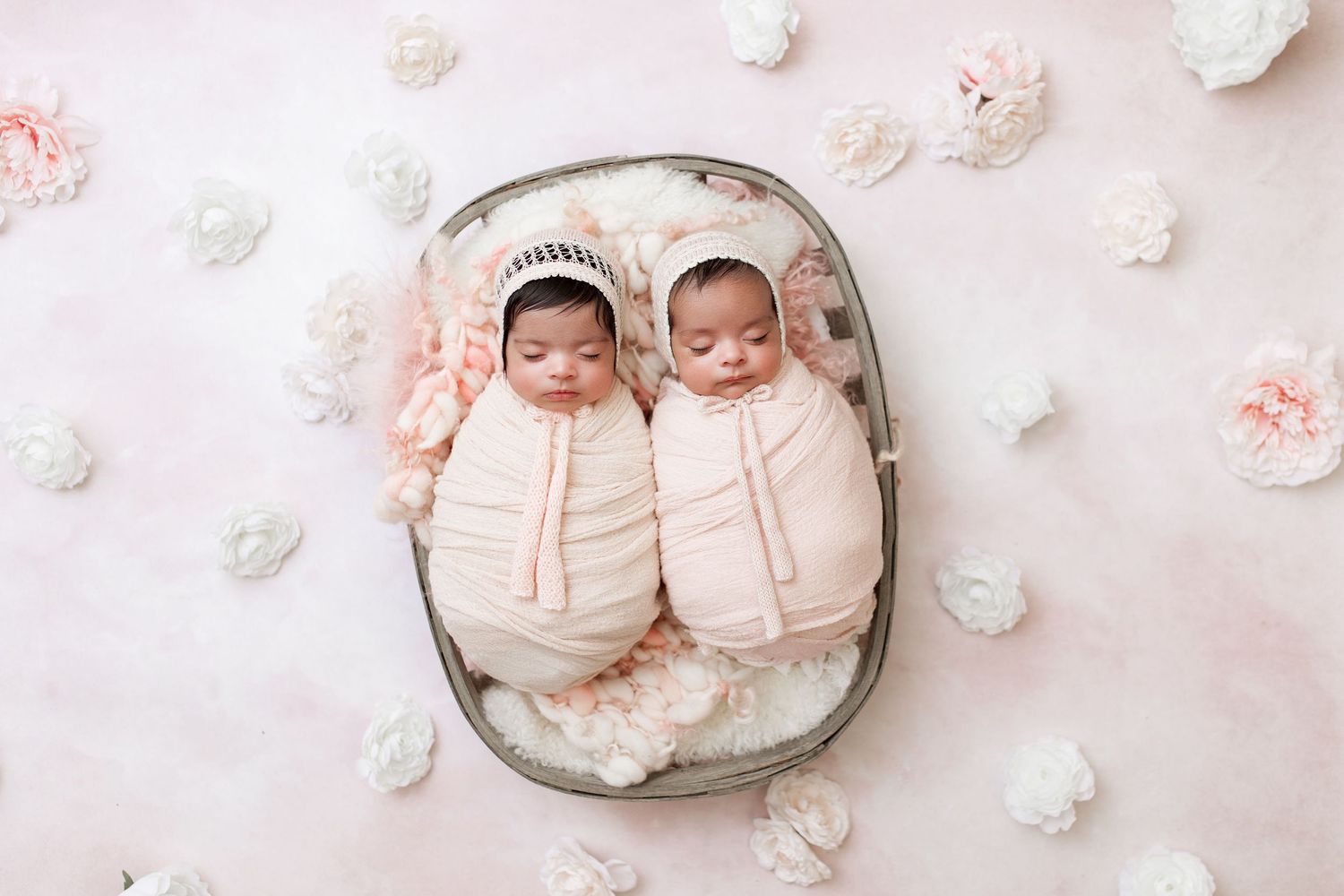 Twin babies in matching outfits sleep side by side in a grey basket surrounded by pink and white flowers.