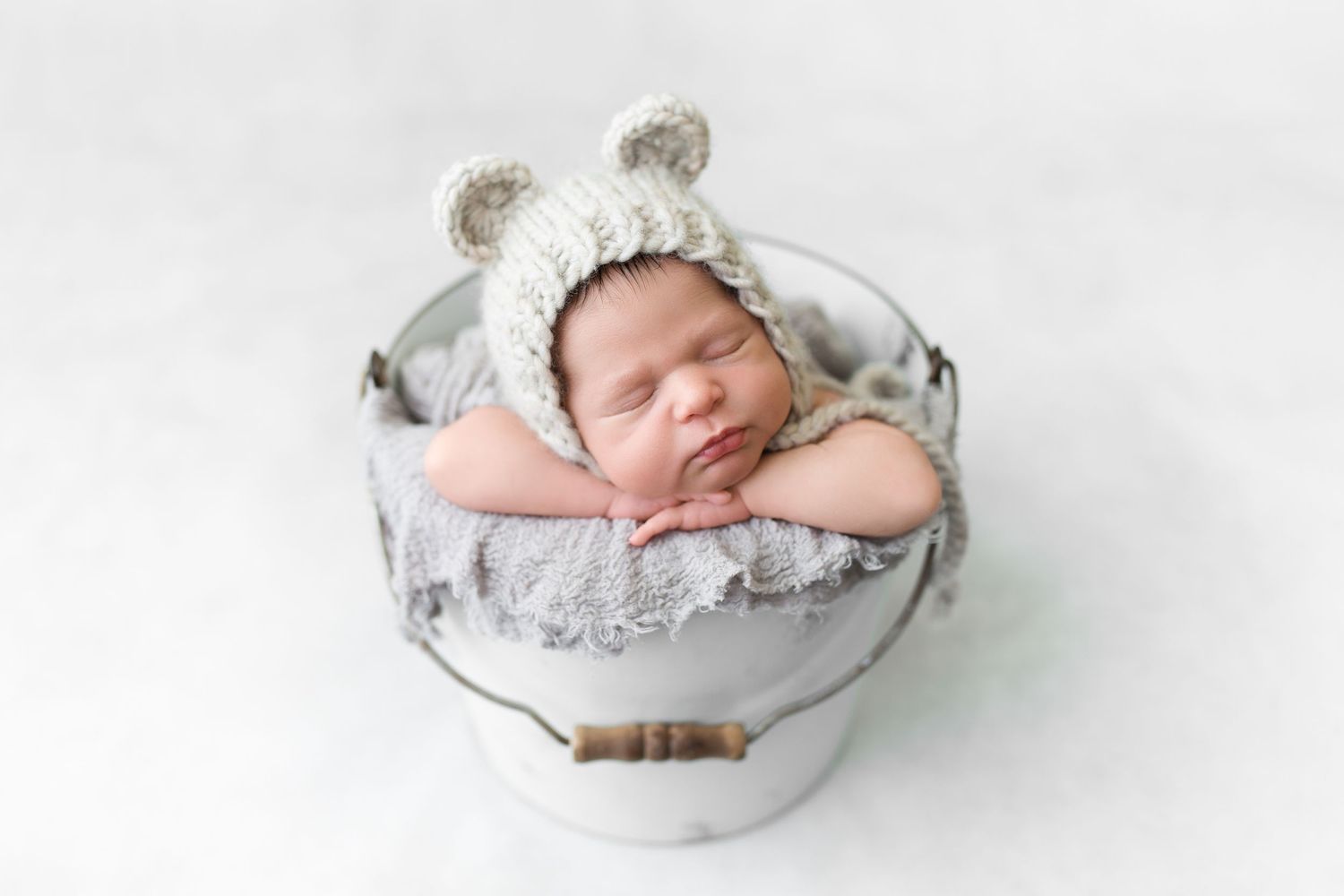 Sleeping newborn wearing bear ear bonnet nestled in decorative gray and white bowl.