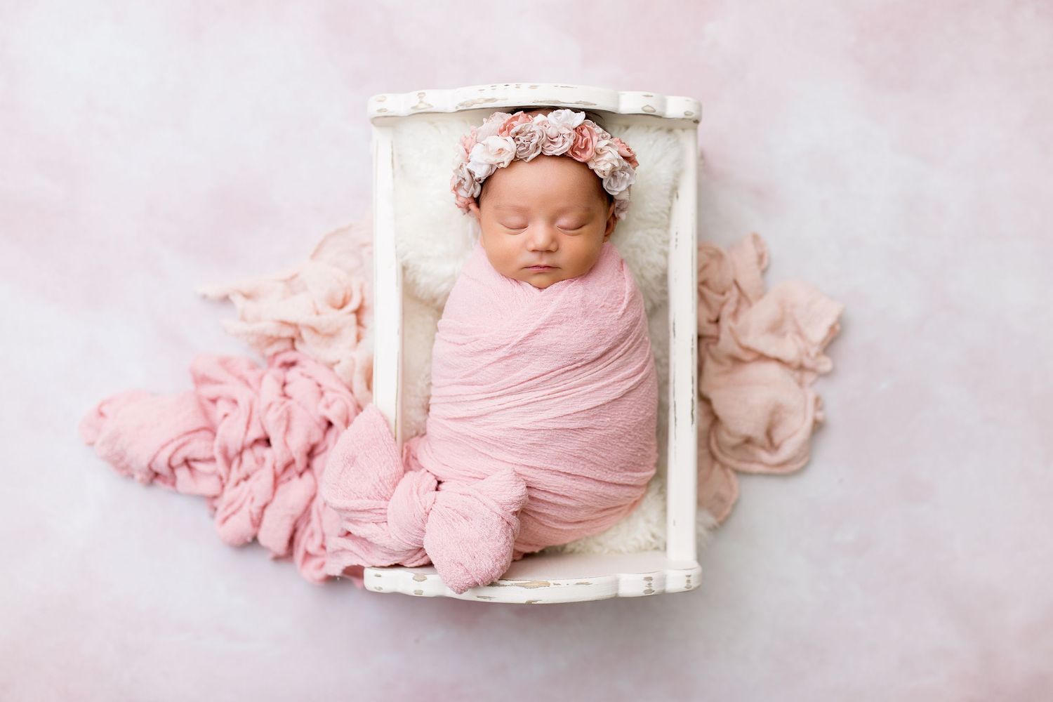 Sleeping newborn wrapped in pink swaddle lying in small white bed with delicate lace details.