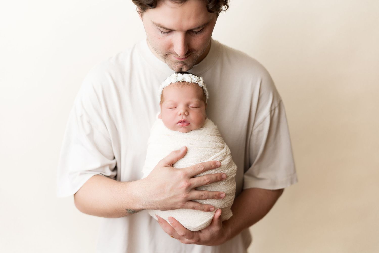 Parent in white shirt tenderly holds sleeping newborn baby wrapped in white against neutral backdrop.
