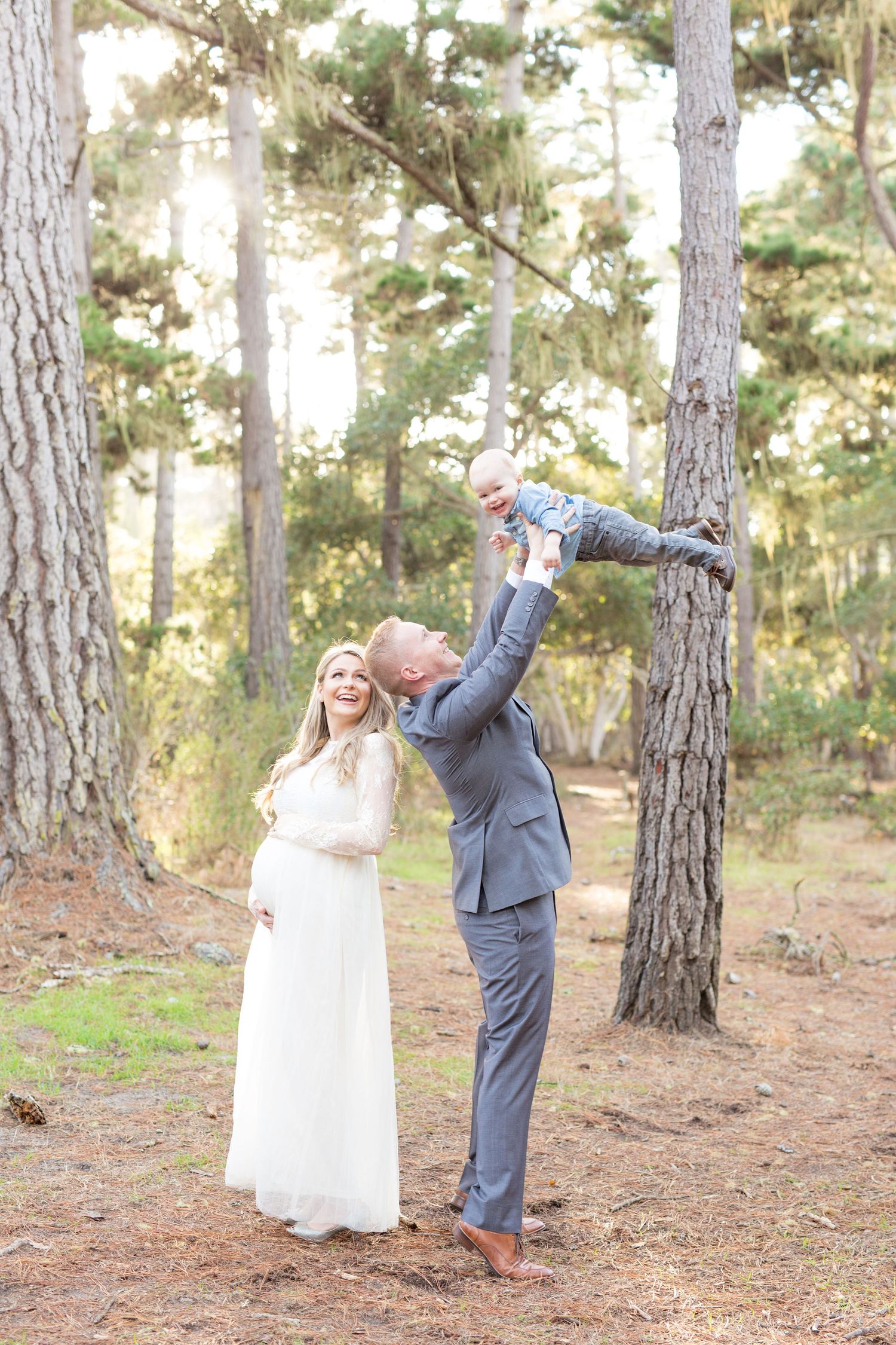 A playful couple shares joyful moments between pine trees in a sunlit forest.