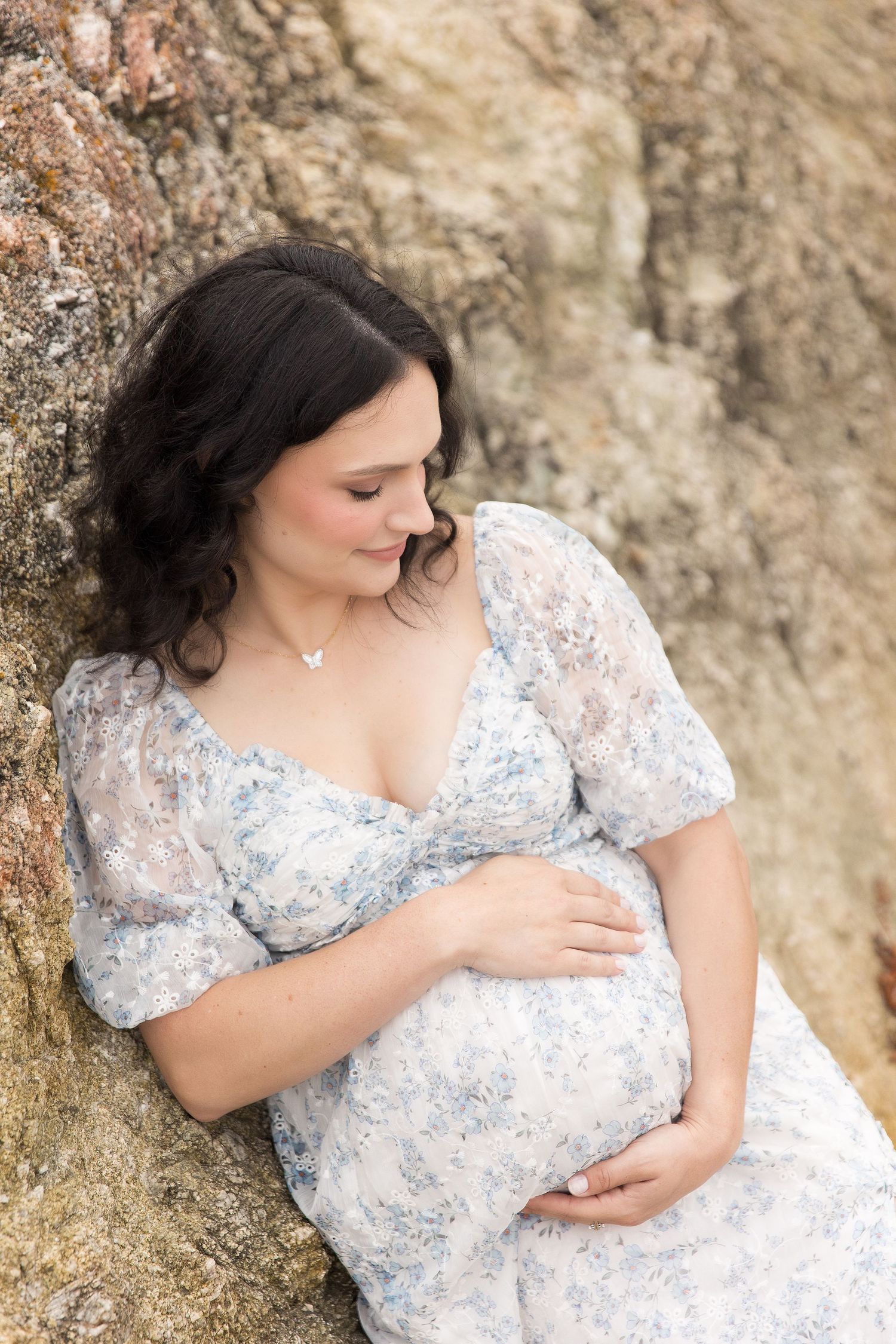 Elegant maternity portrait against textured rock wall in delicate floral dress.