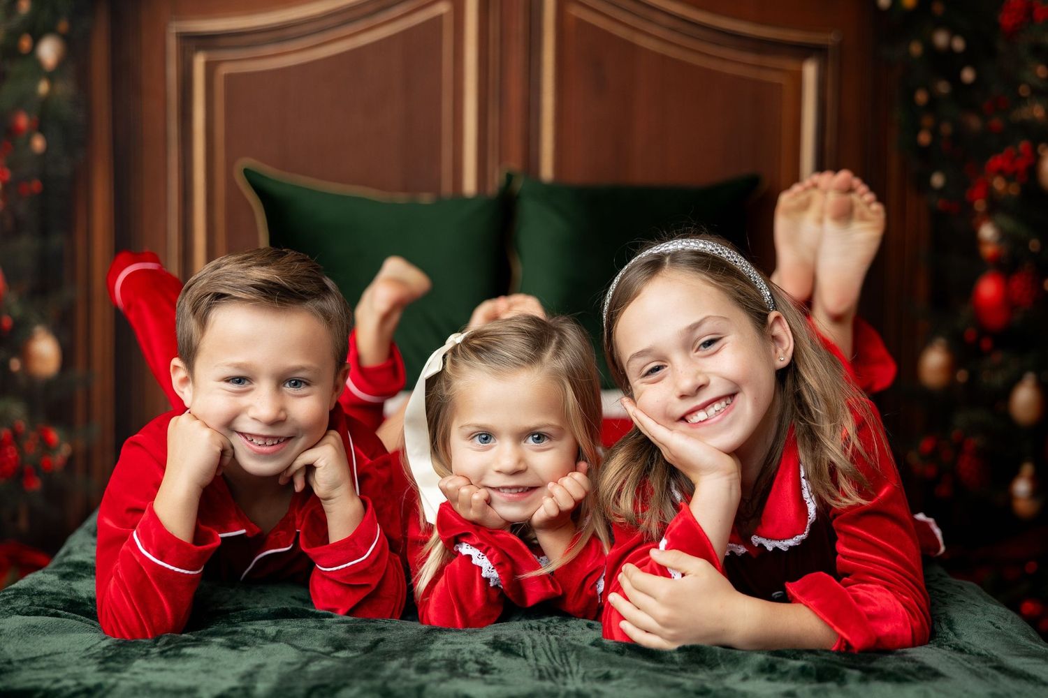 Three siblings in red Christmas pajamas lying on a green holiday bed at a Milk & Cookies mini session in Pacific Grove