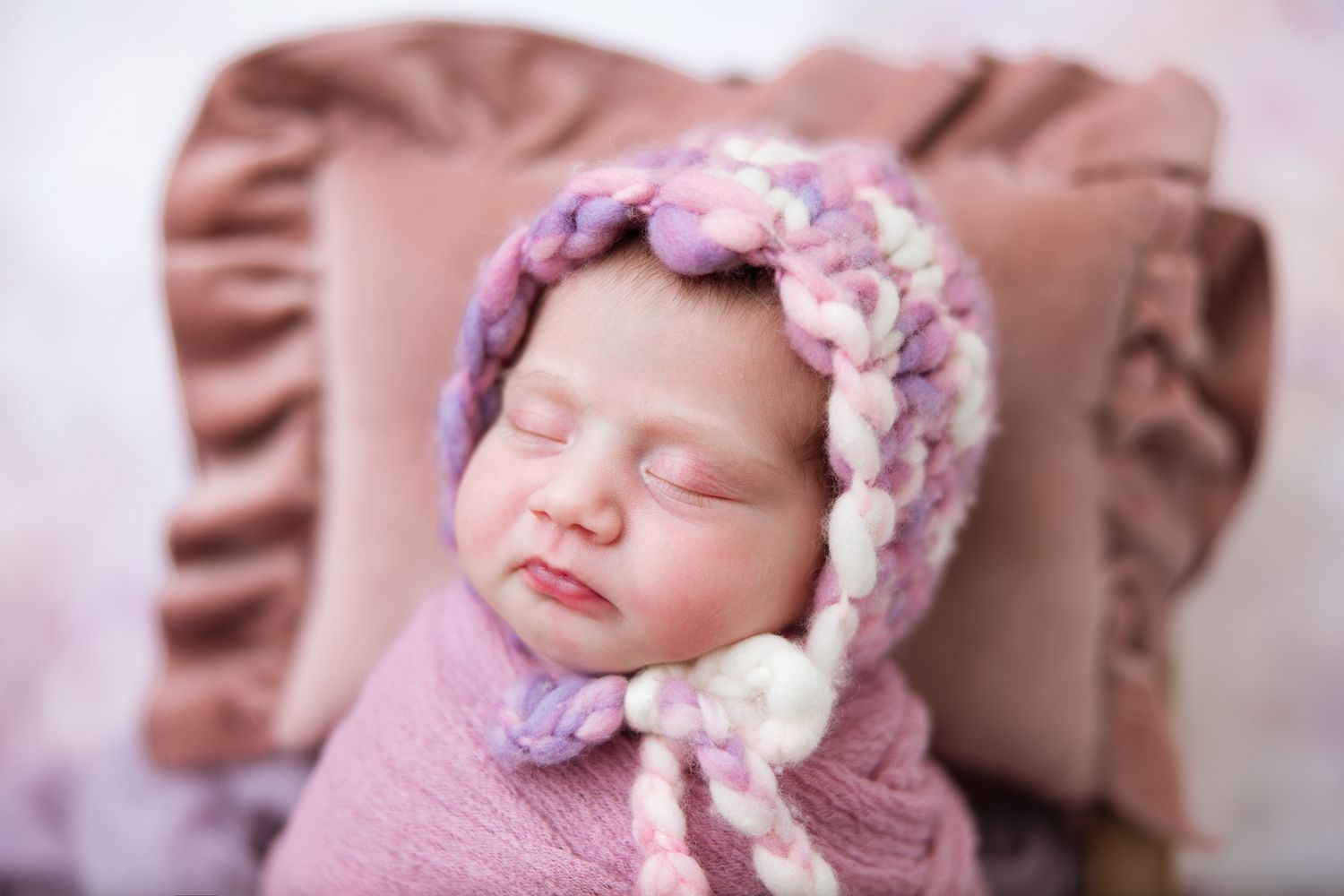 A sleeping newborn baby wearing a delicate pink floral bonnet rests peacefully on soft pink fabric.