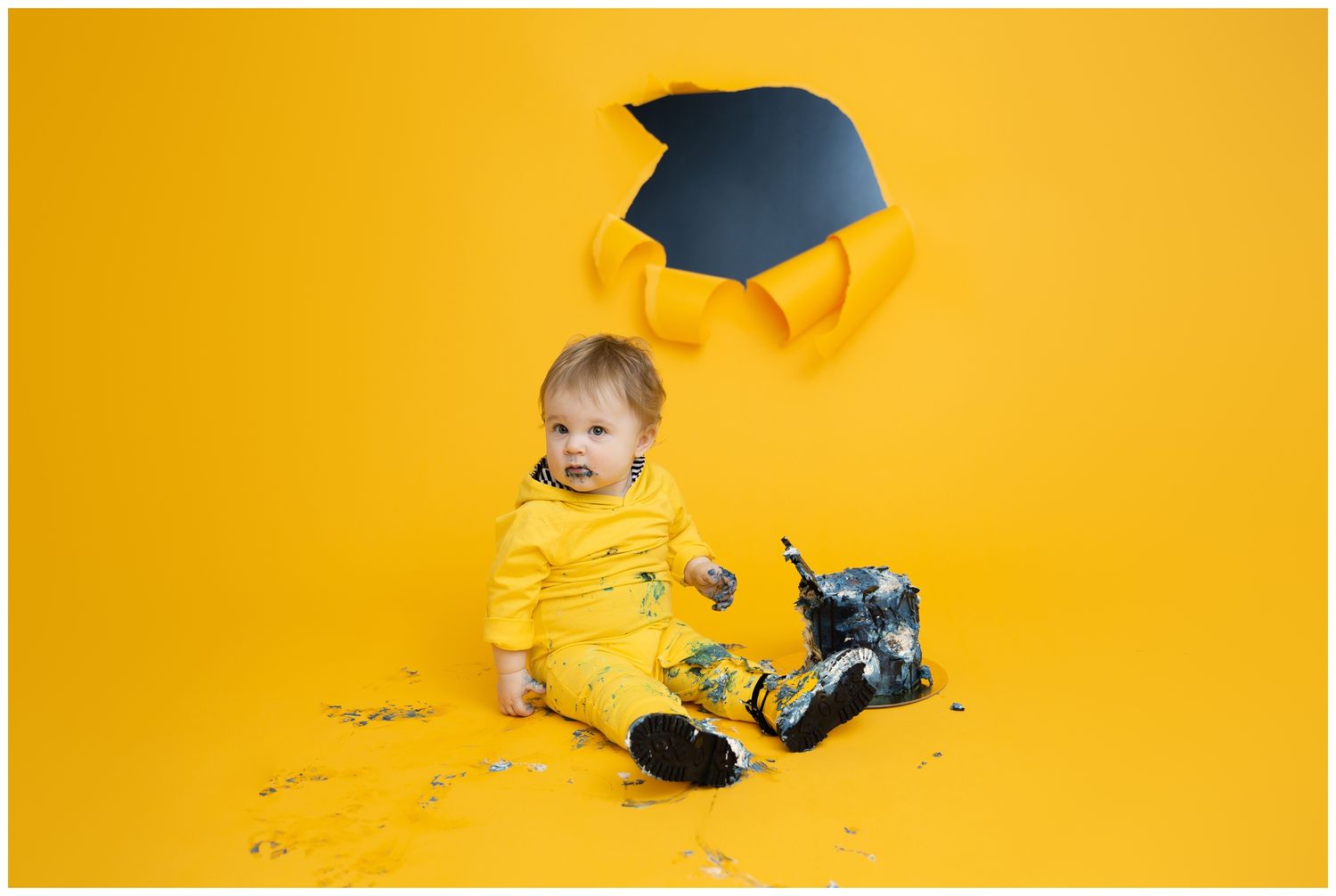 Small child wearing yellow outfit sits on yellow backdrop with graduation cap and scattered money around.