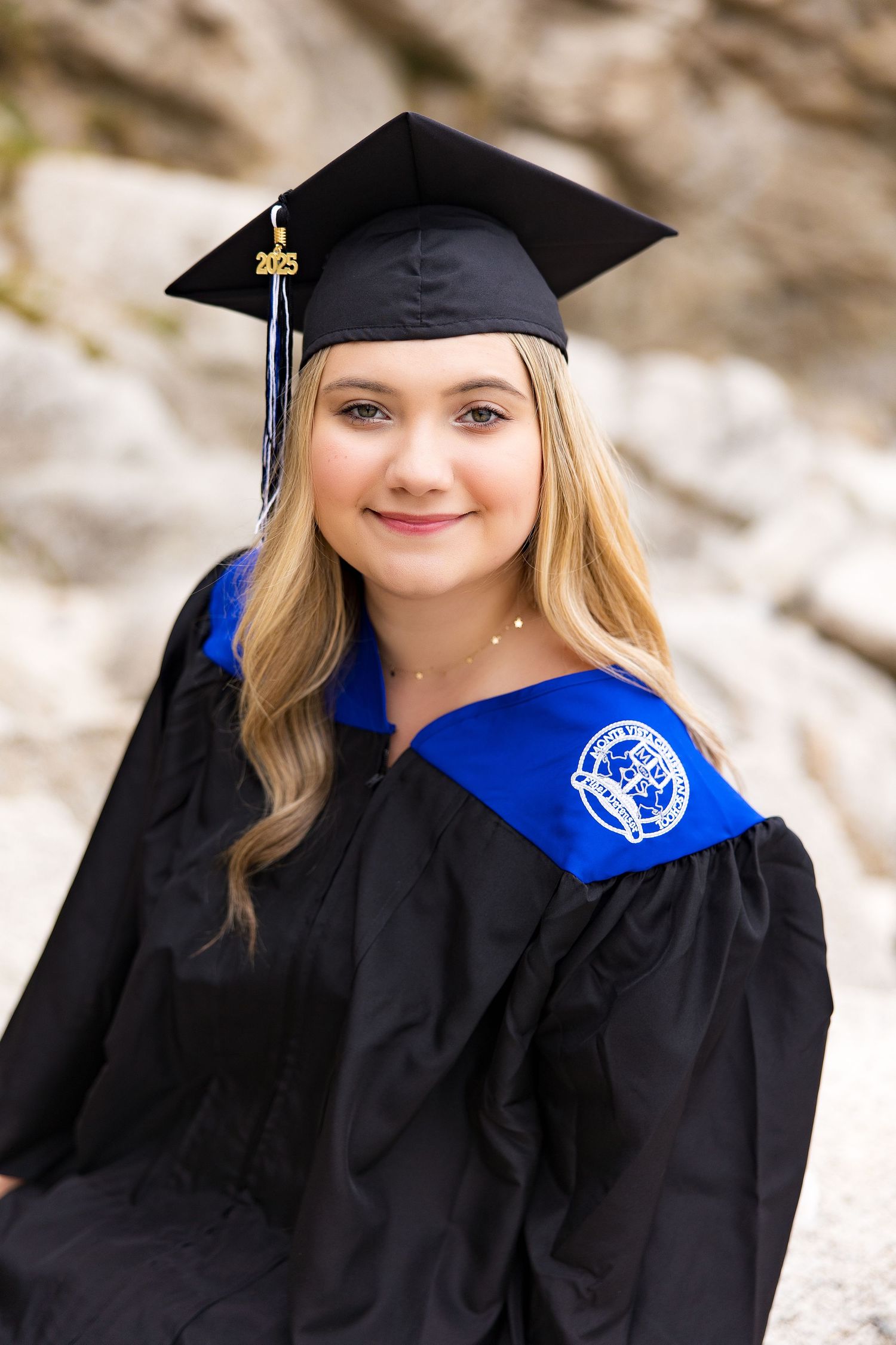 Young woman in black graduation cap and gown with blue hood smiles outdoors against rocky background.