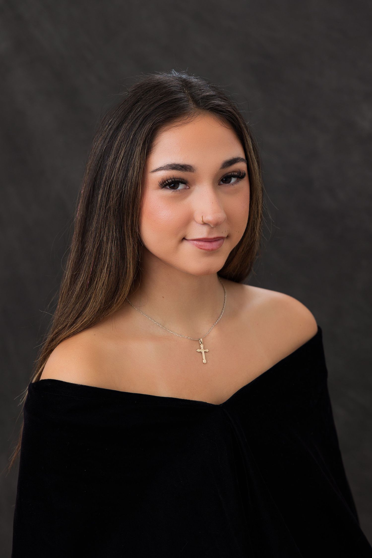 Young woman with long brown hair wearing a black off-shoulder top and gold cross necklace poses for a portrait.