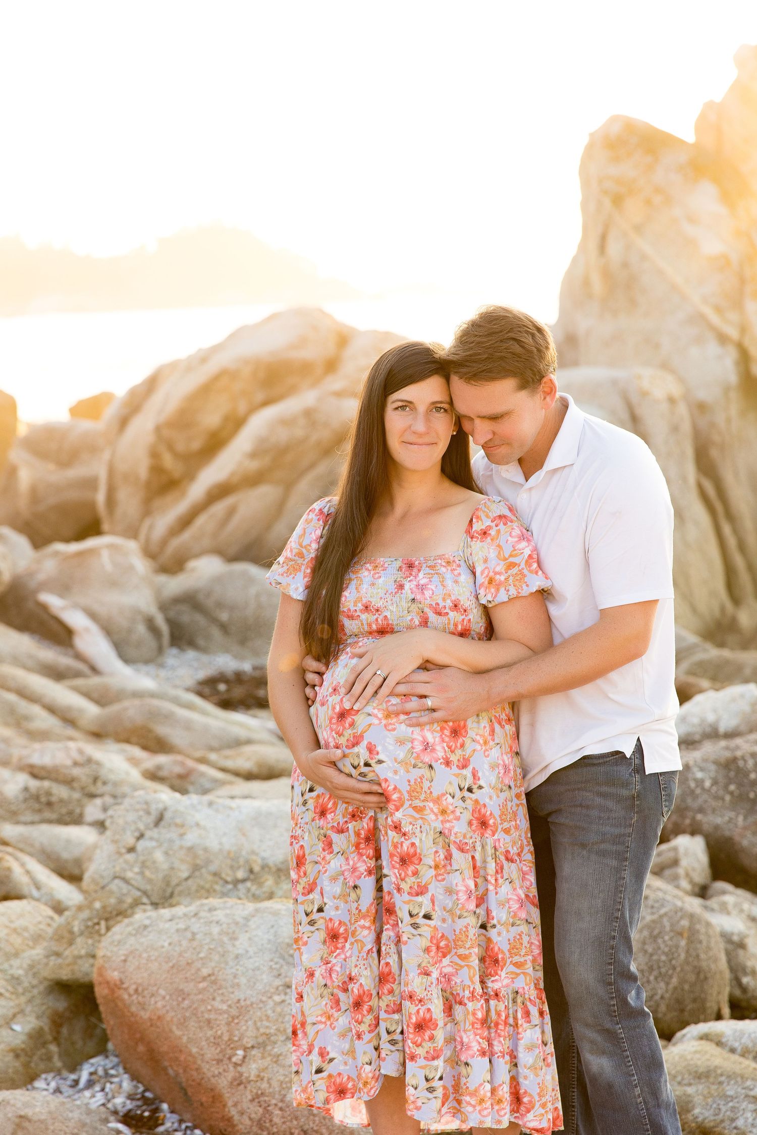 Couple embraces on rocky beach during sunset while wearing floral dress and white shirt.