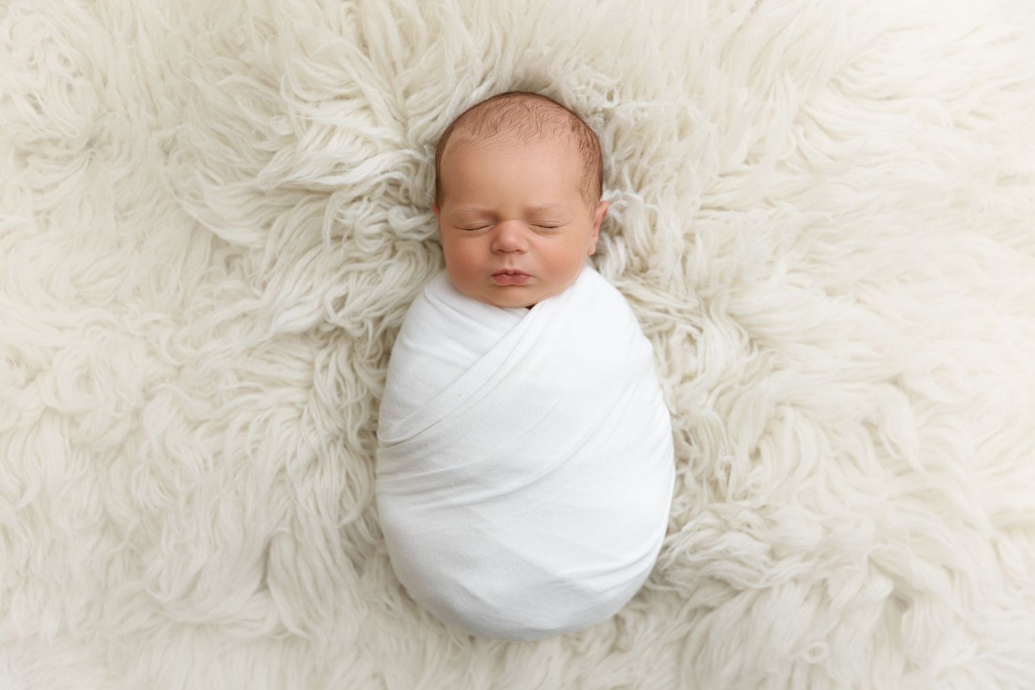 A sleeping newborn baby swaddled in white fabric lies peacefully on a soft furry white blanket.