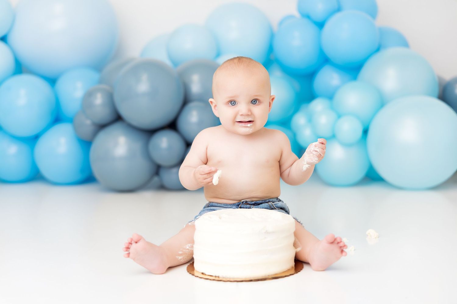 Baby in white diaper poses with cake for first birthday photo shoot against blue balloon background.