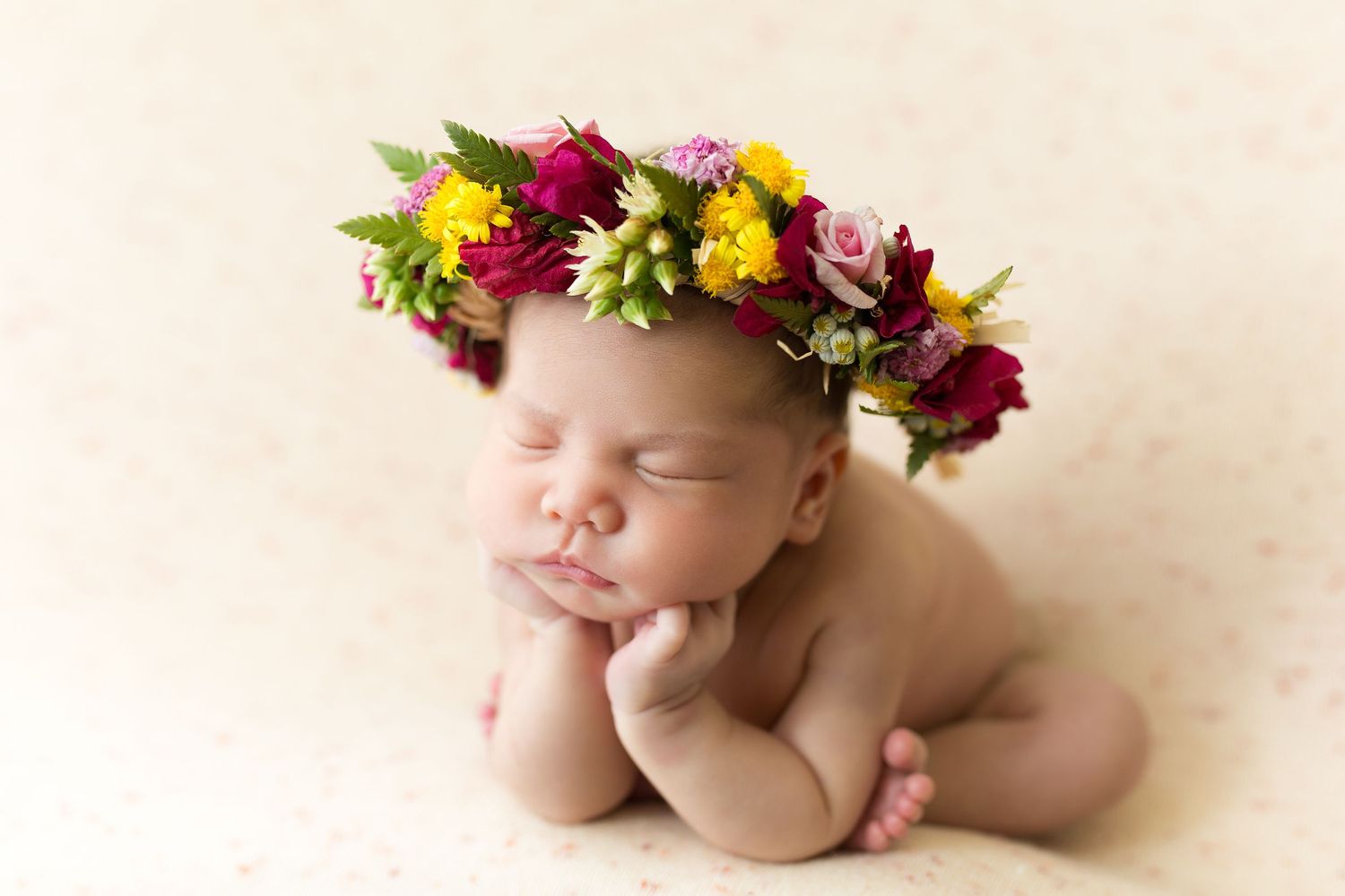 A sleeping baby wearing a colorful floral crown lies peacefully on a cream-colored background.