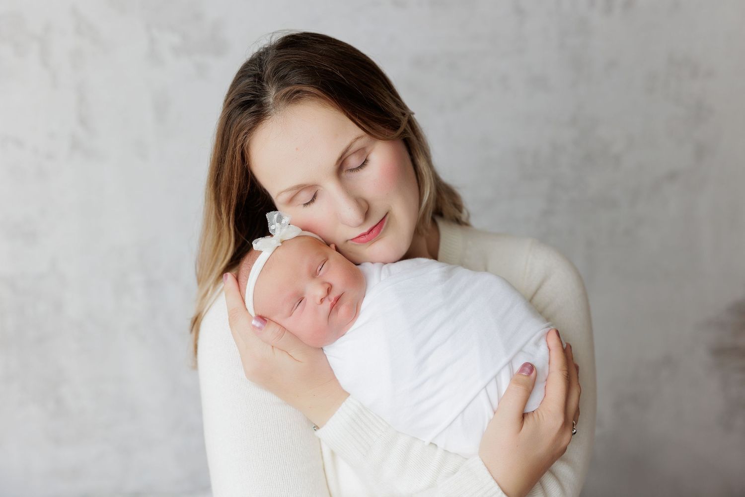 Mother tenderly holding sleeping newborn baby wrapped in white swaddle against gray background.