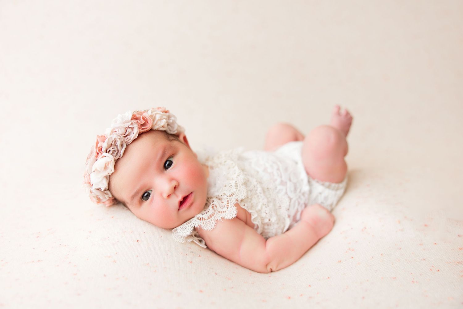 Baby in lace romper and floral crown lying on cream colored blanket.