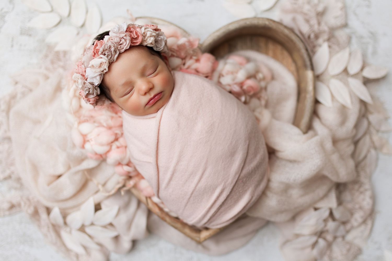 Sleeping newborn in pink wrap with floral headband in curved basket.