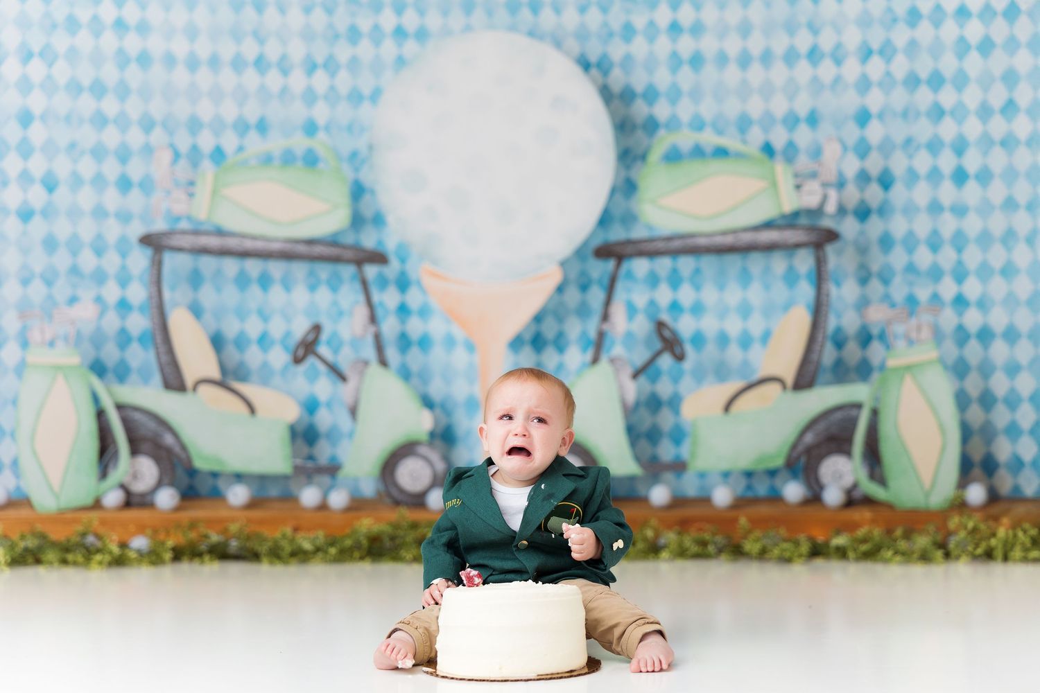 Baby smiling next to white cake against blue checkered background during cake smash photo session.