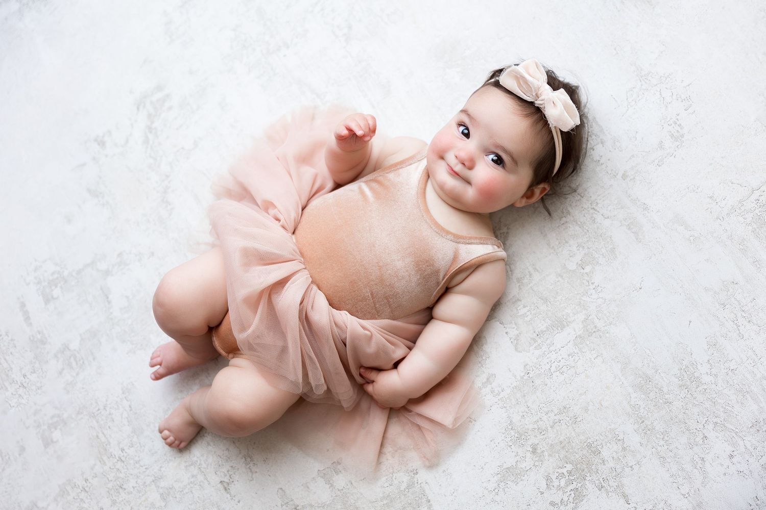 Baby wearing pink tulle outfit and bow lies on white floor during portrait session.