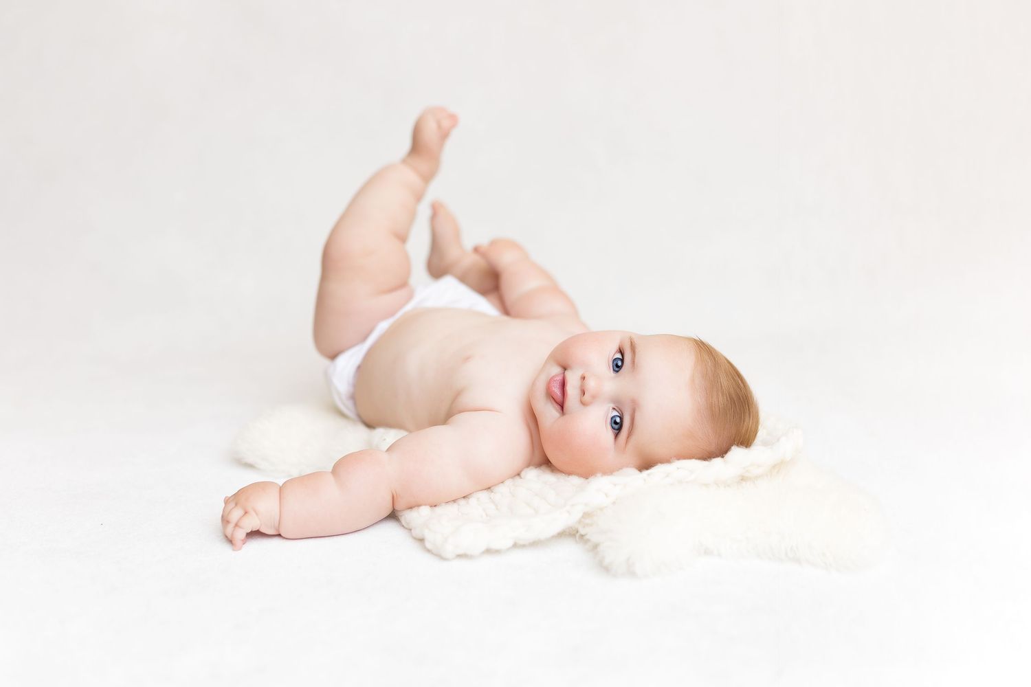 Happy baby in white diaper lying on soft white blanket.