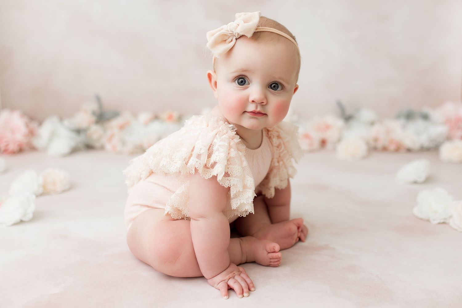 Baby in cream lace outfit on pink backdrop with scattered floral decorations.
