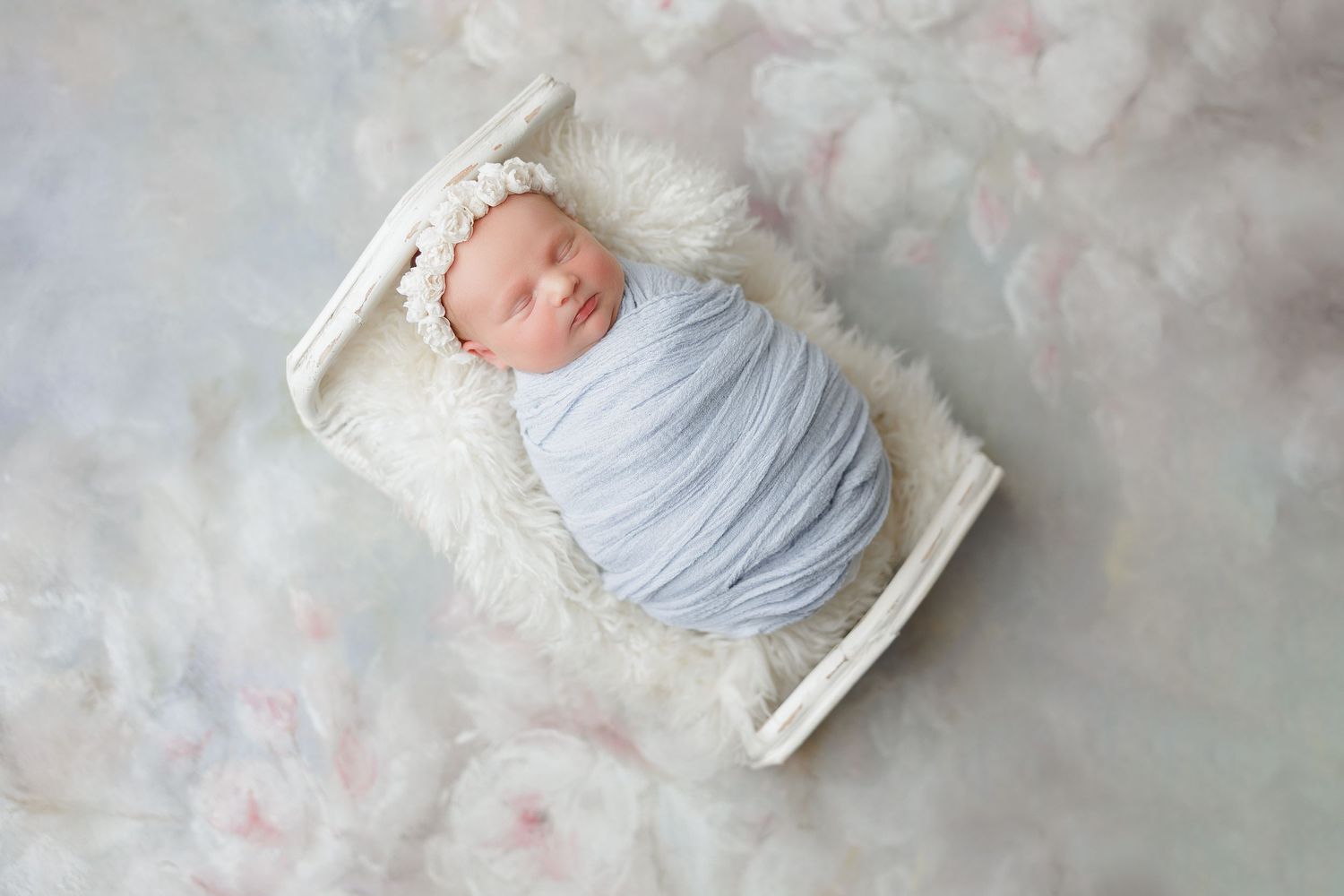 Newborn baby swaddled in light blue wrap sleeping in white mini crib on floral backdrop with lace headband.