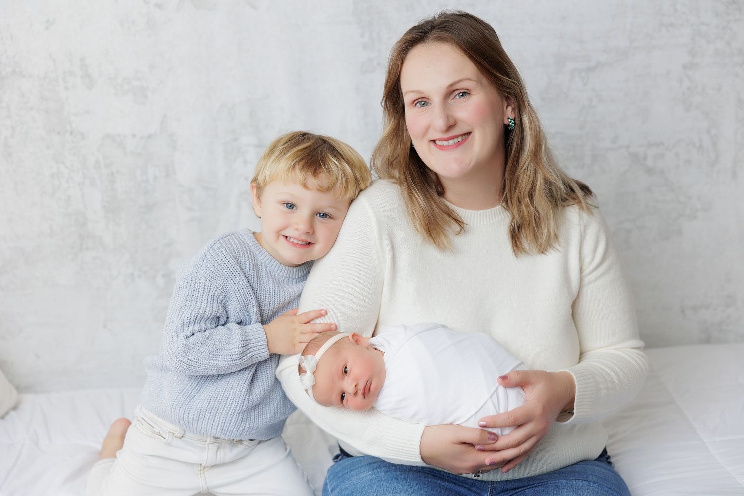 A mother holds her newborn baby while her young toddler son smiles beside her in a bright studio setting.