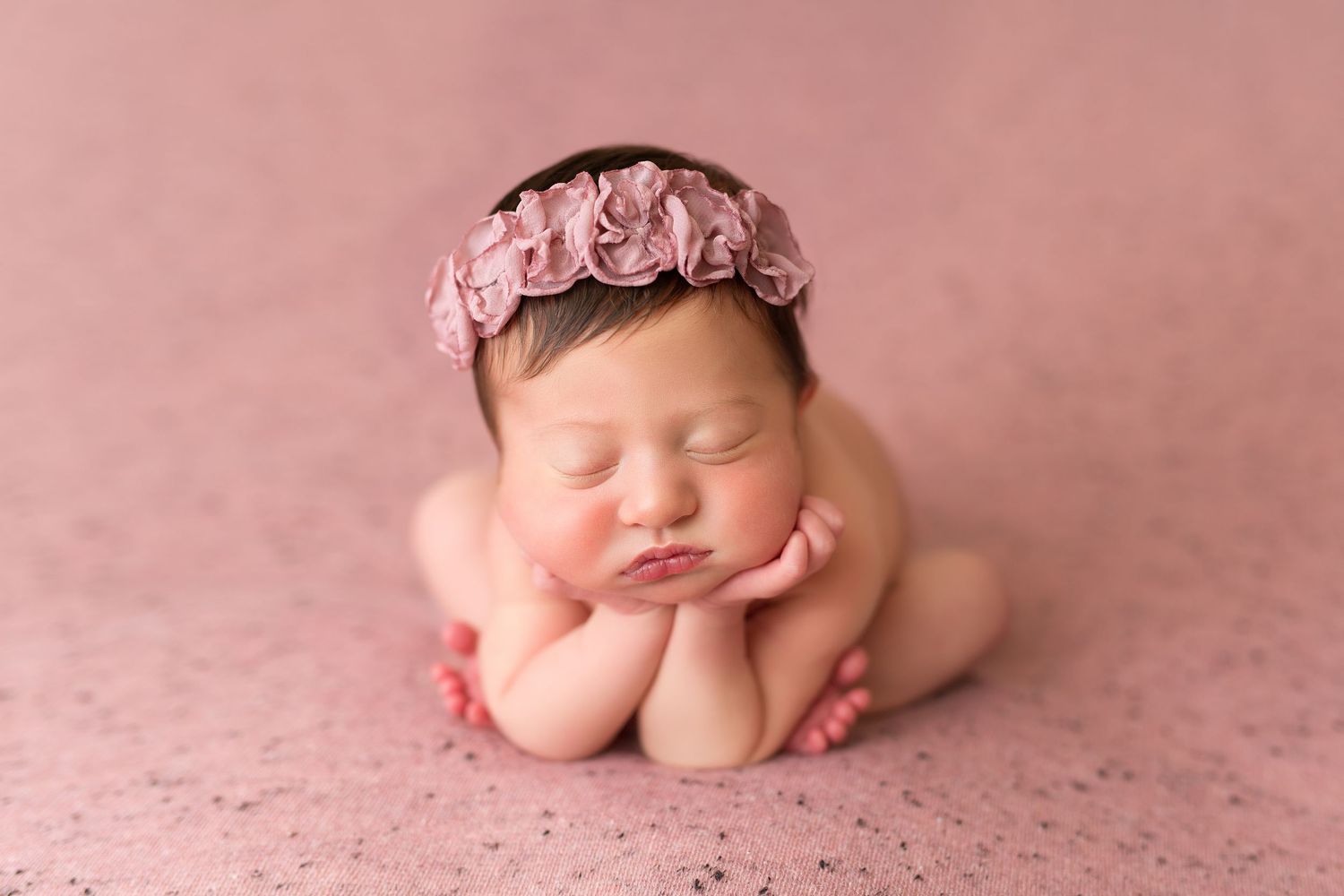 Sleeping newborn posed with head on hands wearing pink floral headband on pink backdrop.