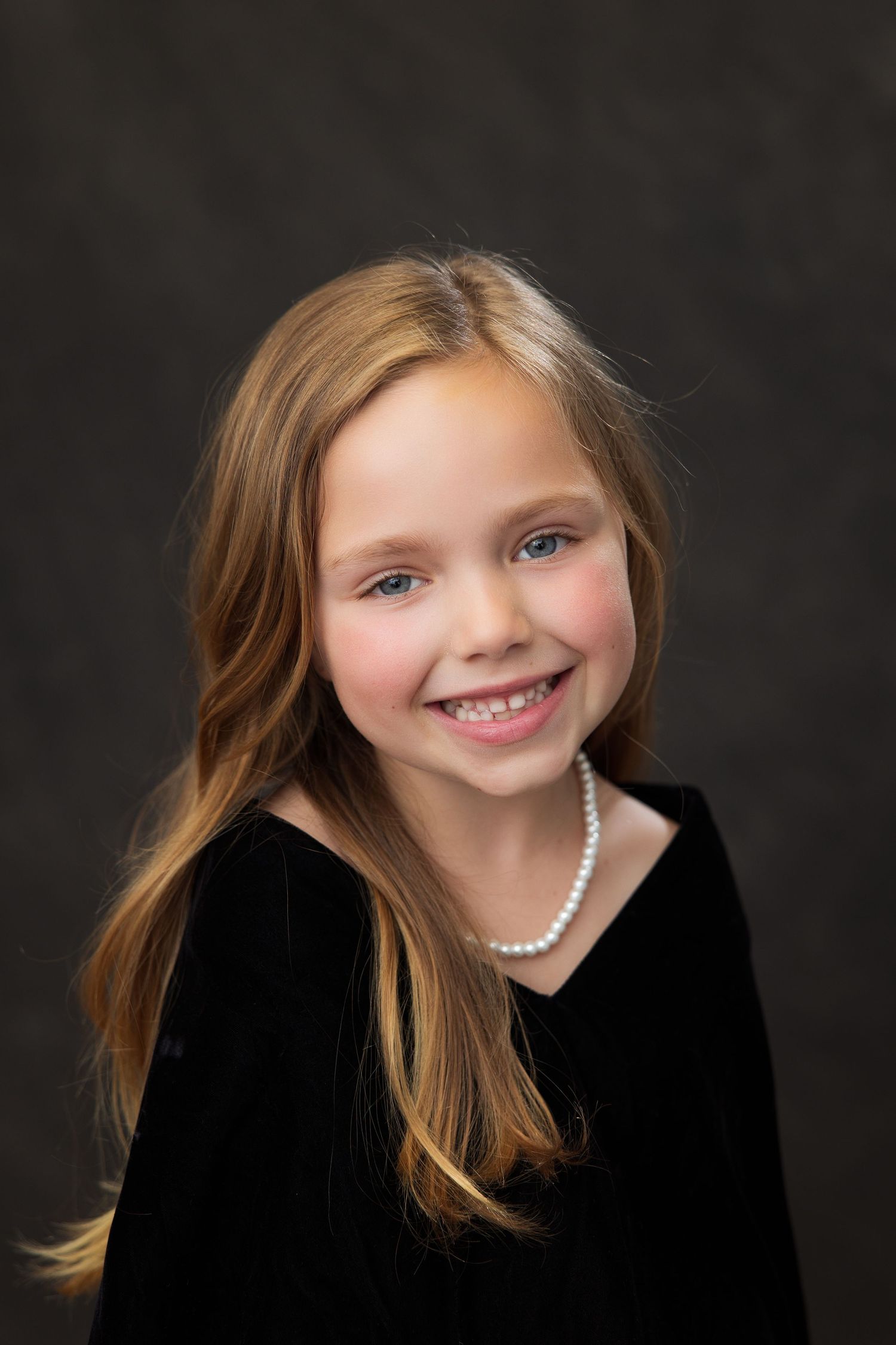 Studio portrait of a young person with brown hair wearing a black sweater and pearl necklace against dark background.