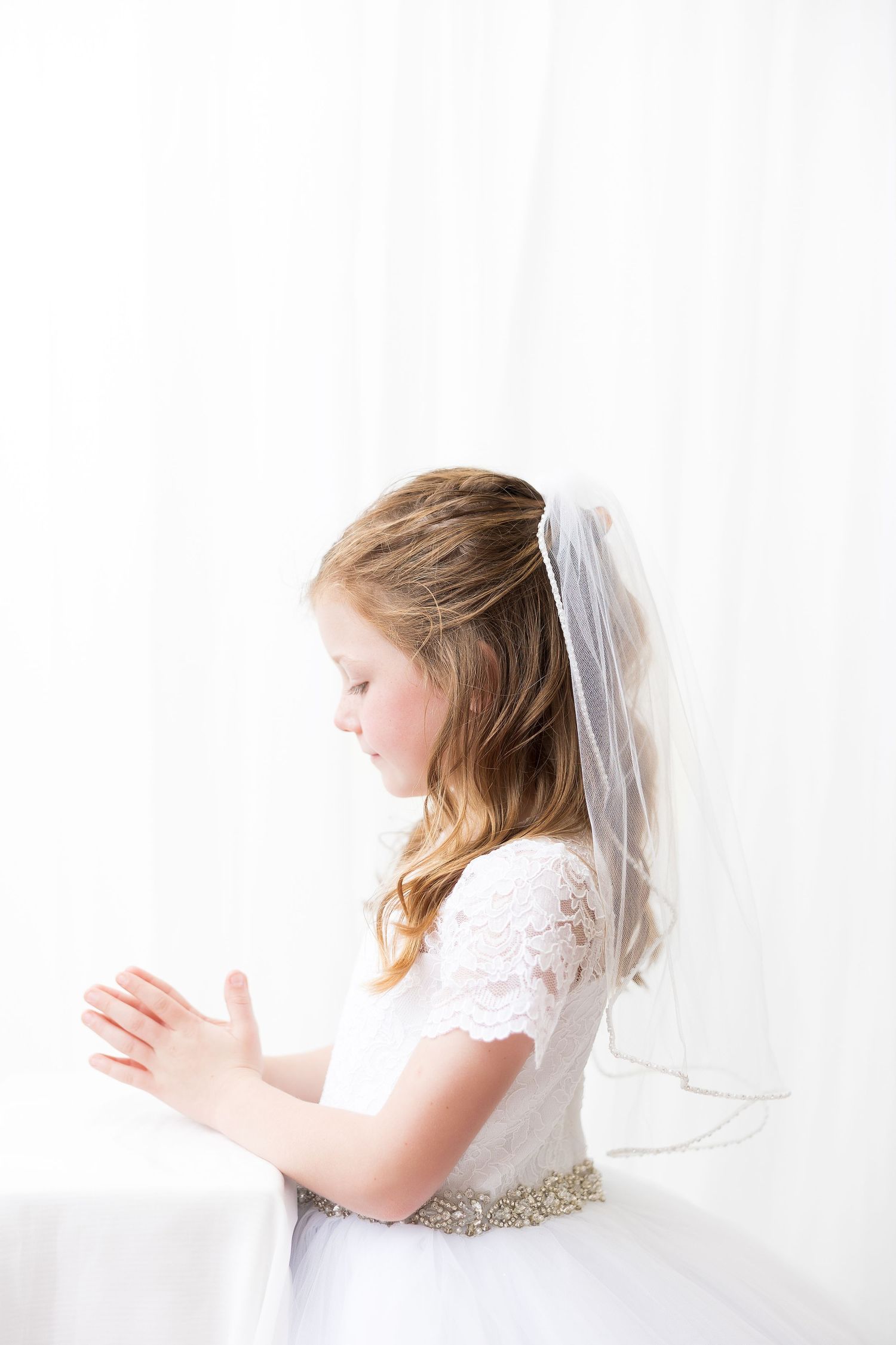 A profile view of a child in white dress with veil praying with hands clasped together.