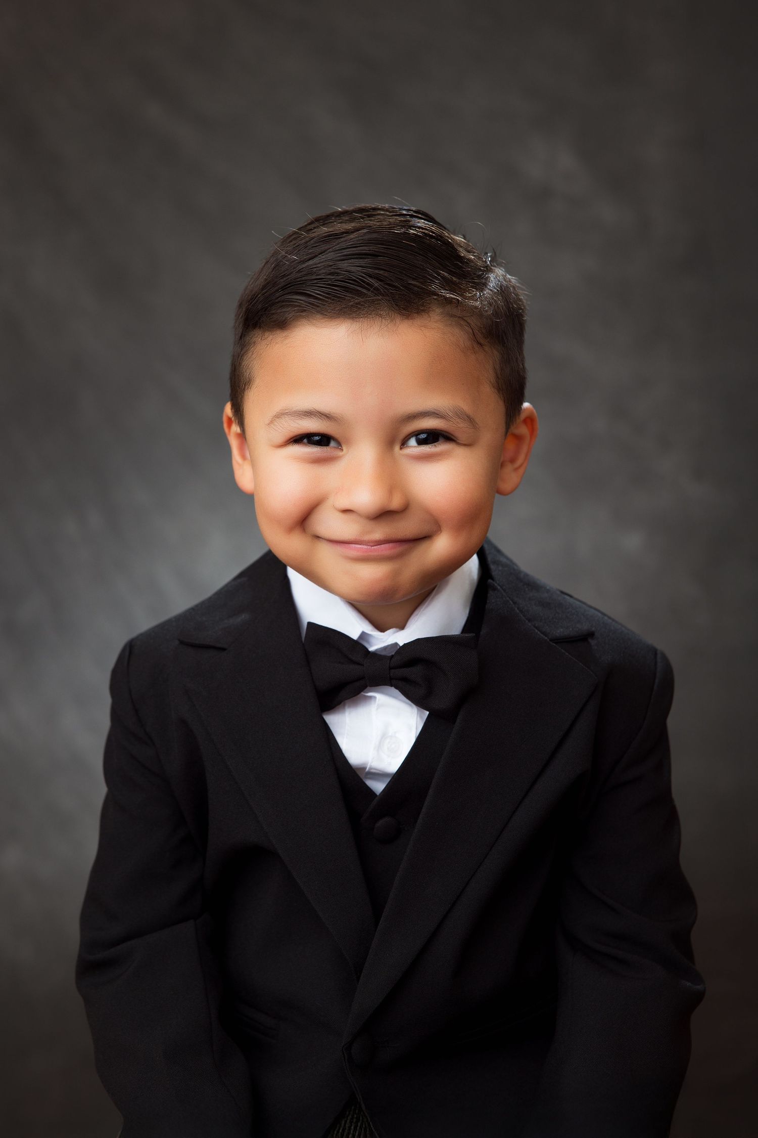 A young child smiles in a formal black tuxedo and bow tie during a studio portrait session against a gray backdrop.