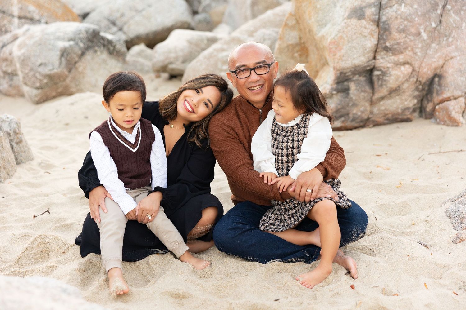 Family sitting close together on sandy beach rocks during portrait session.