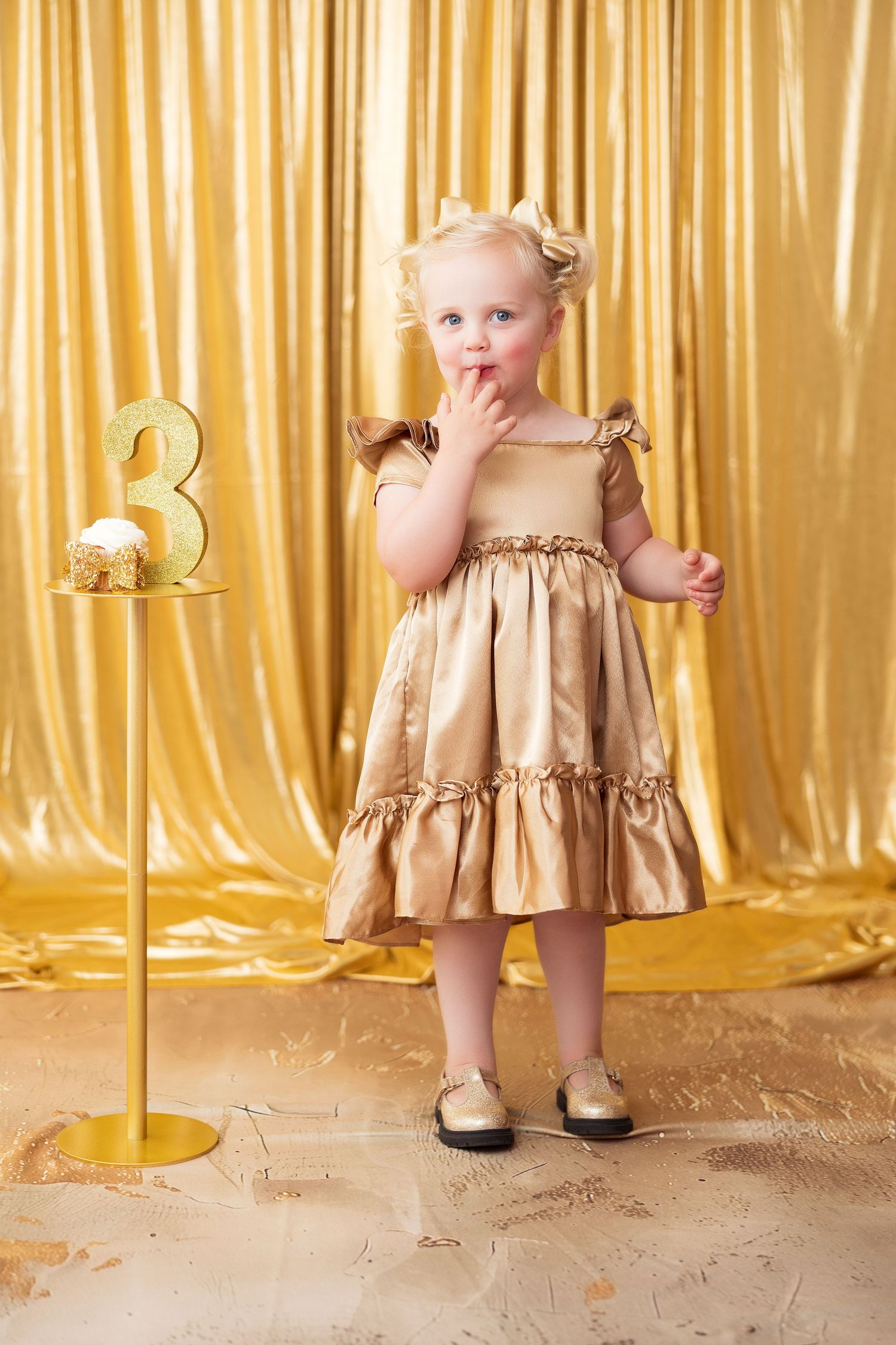 A toddler in a gold dress stands against a shimmering gold curtain backdrop in three similar poses.