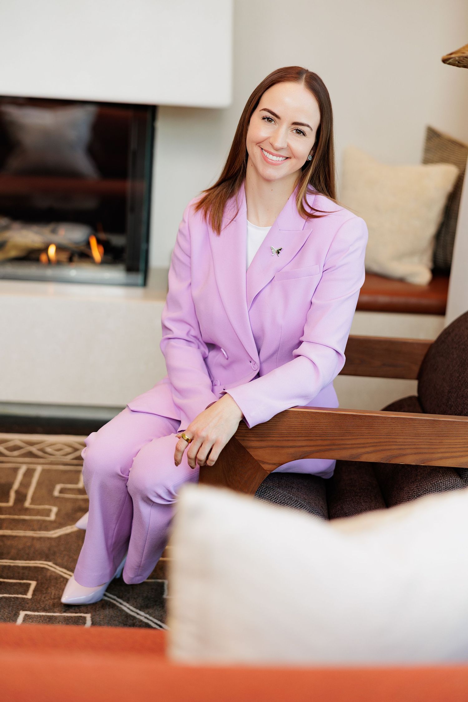 Woman in lavender pantsuit sitting by fireplace, smiling confidently in modern living room.