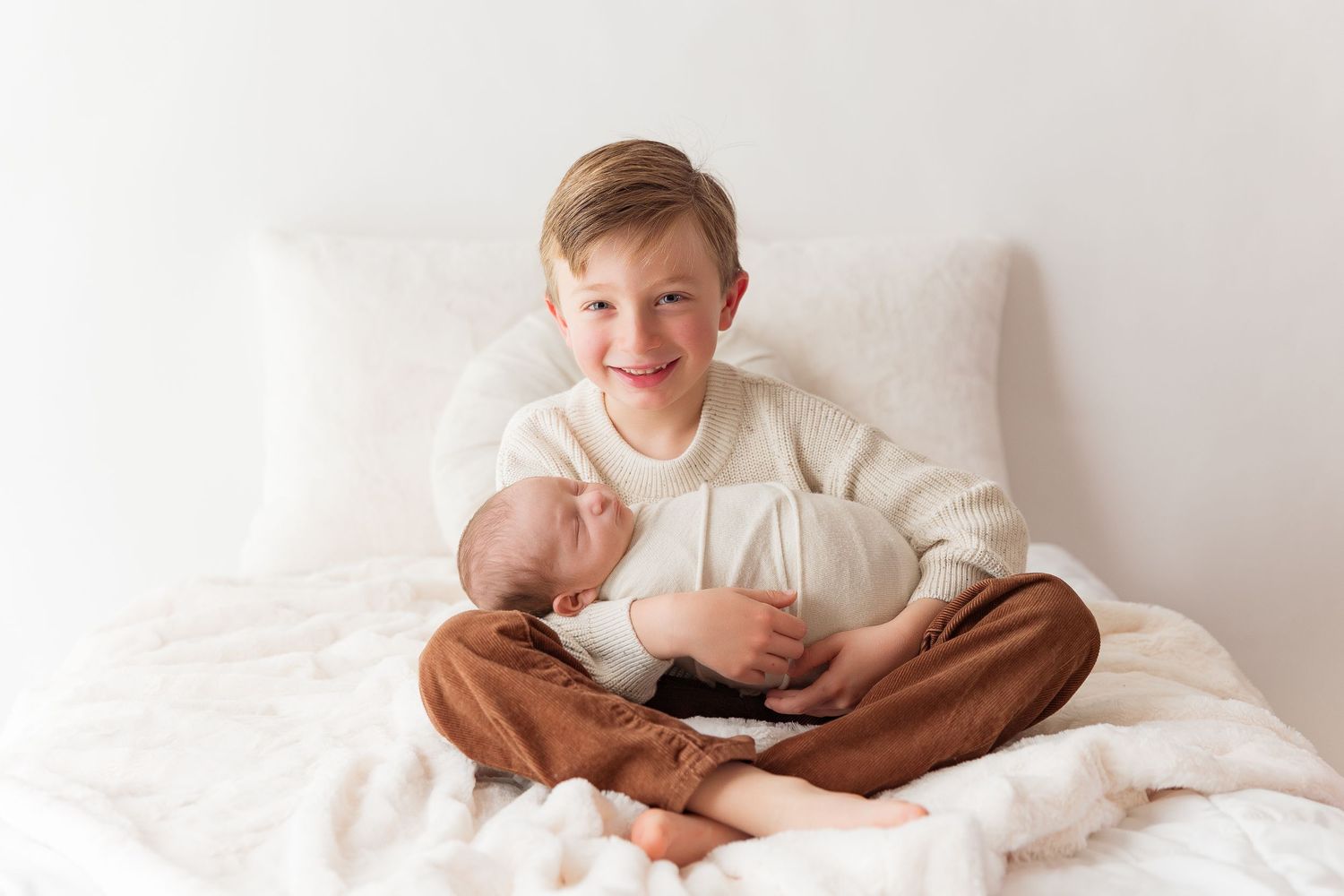 Young boy in white sweater holds sleeping infant on white bedding.