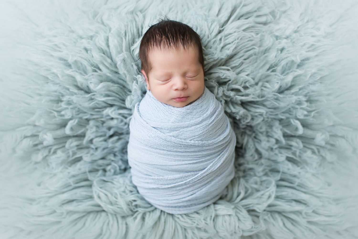 Newborn wrapped in grey fabric centered on circular light blue textured blanket.