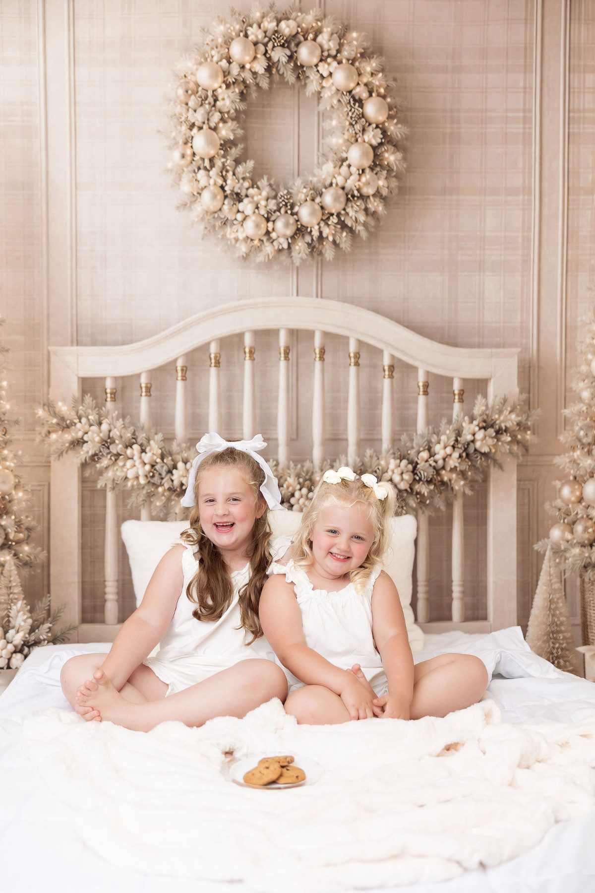 Sisters in white pajamas on a cream Christmas bed set with a gold wreath, Milk and Cookies mini session Monterey