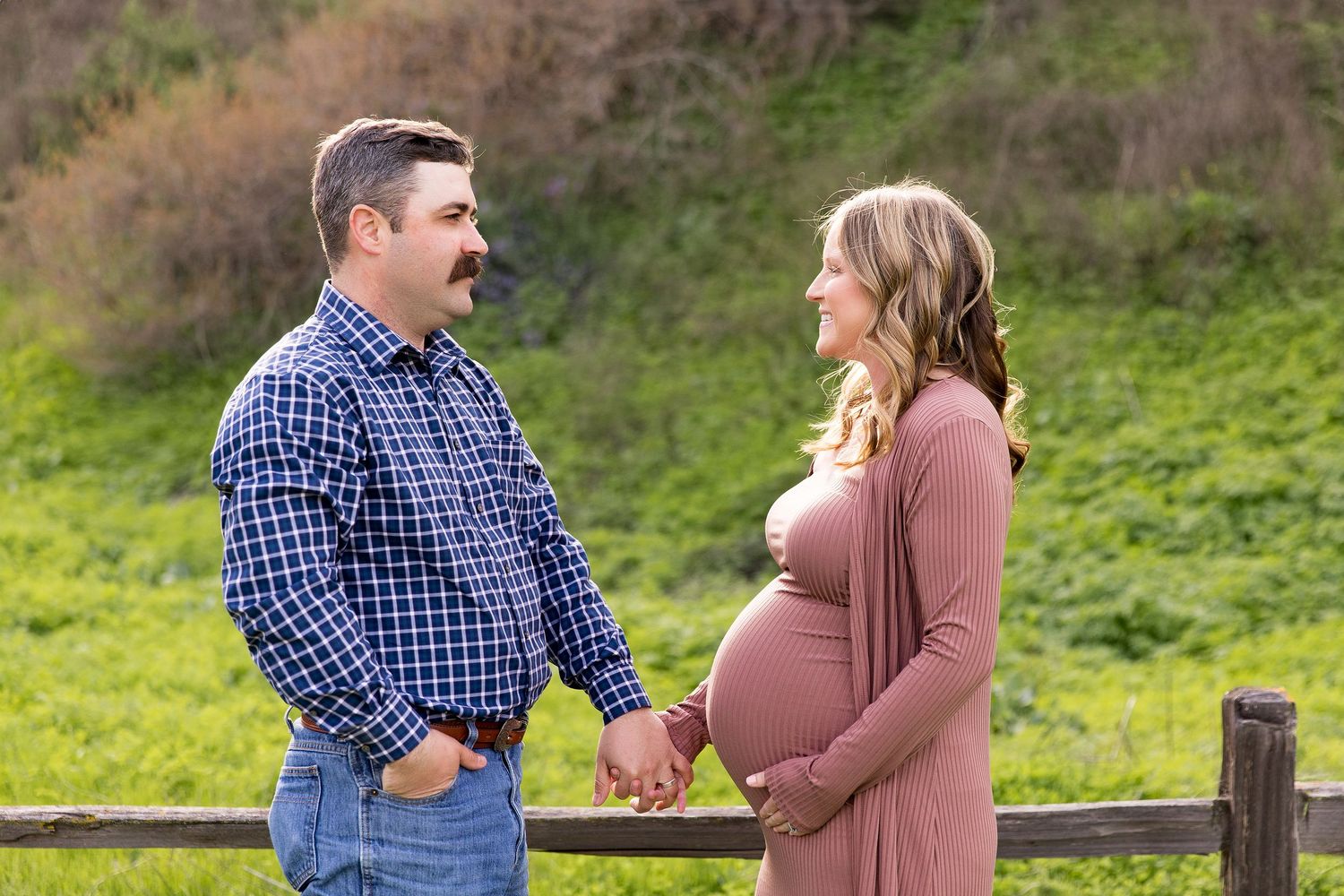 Expecting couple holding hands by a wooden fence in green rolling hills during a Hollister maternity session.