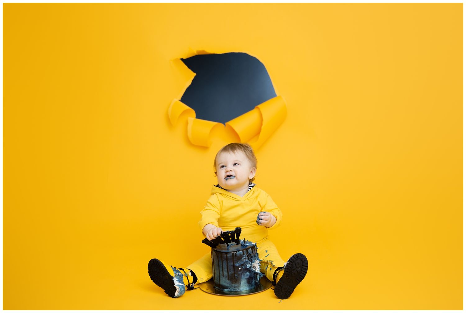 Graduate in black cap and gown sitting cross-legged against bright yellow background.