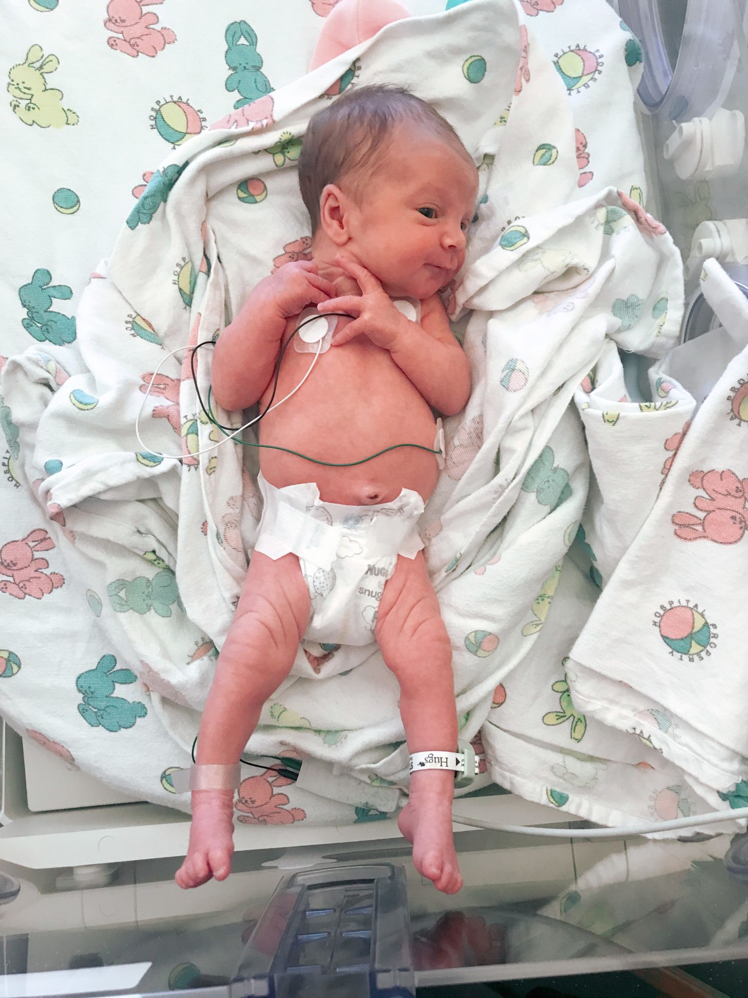 A newborn baby lying on a hospital bed covered with a dinosaur-print blanket.