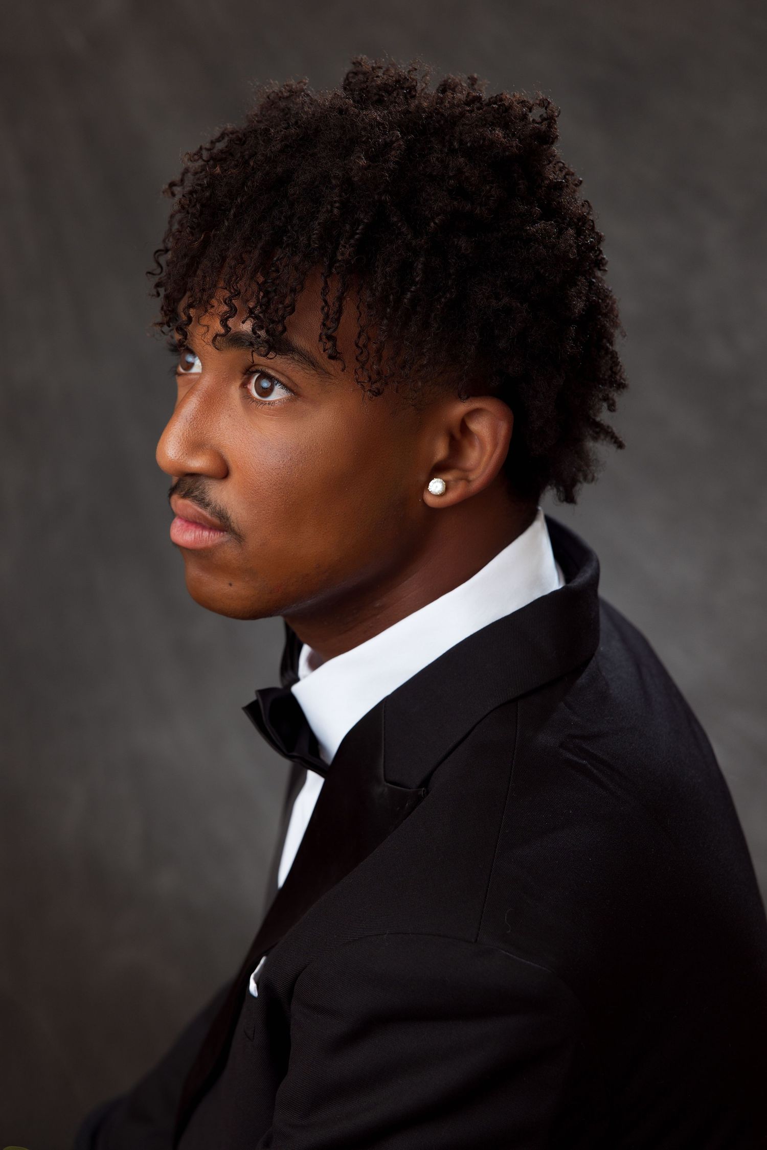 Young Black man in tuxedo with curly hair, mustache, and pearl earring poses against gray background.