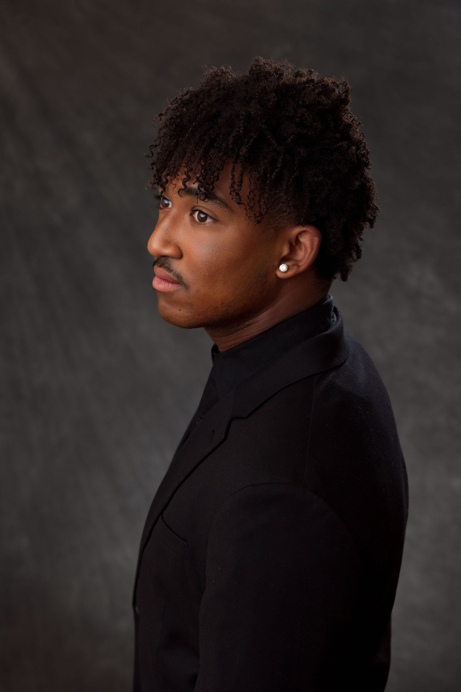 Young Black man in black turtleneck blazer with curly hair and stud earring poses against dark studio backdrop.