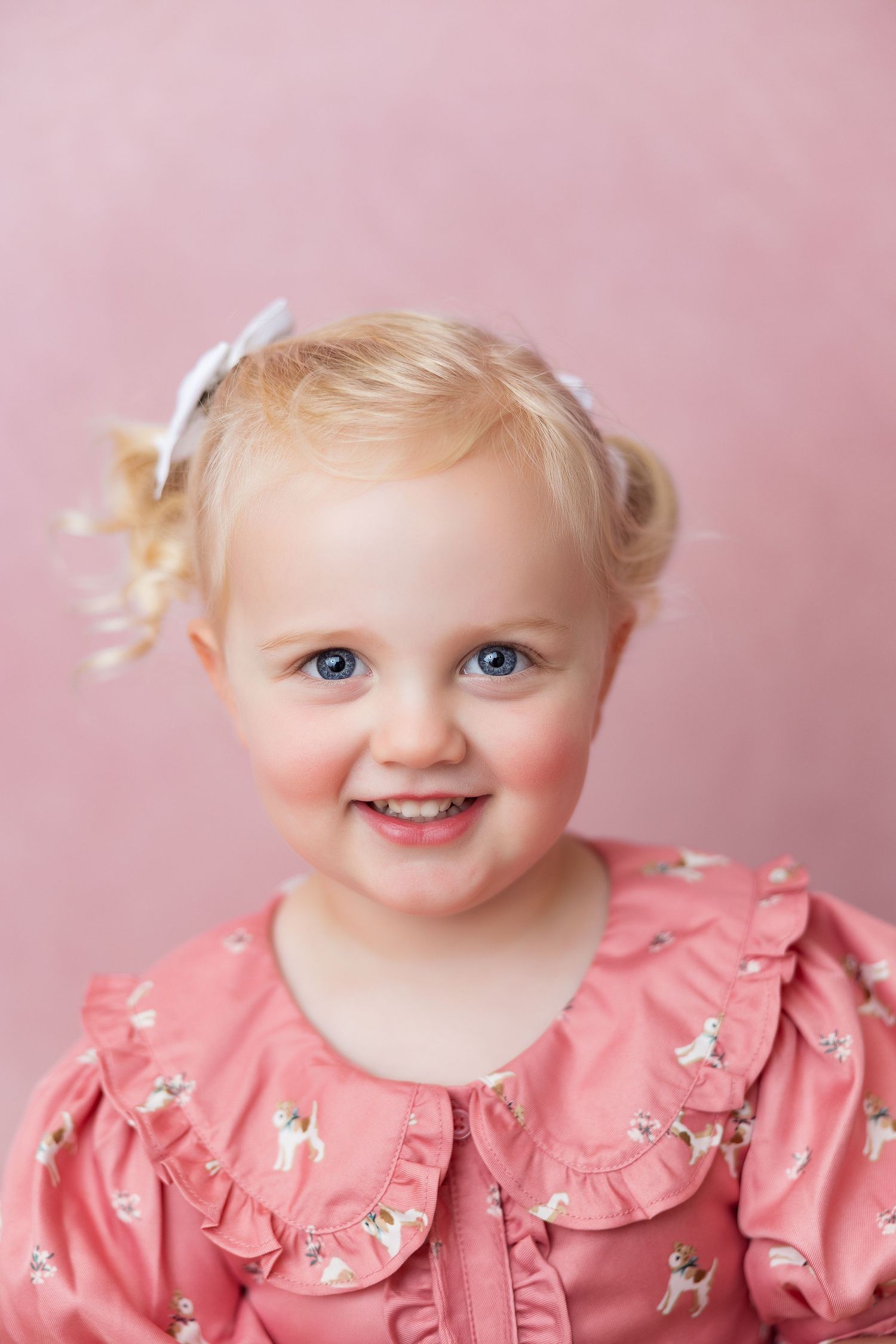 A small child wearing a pink ruffled dress with floral details poses against a soft pink background.