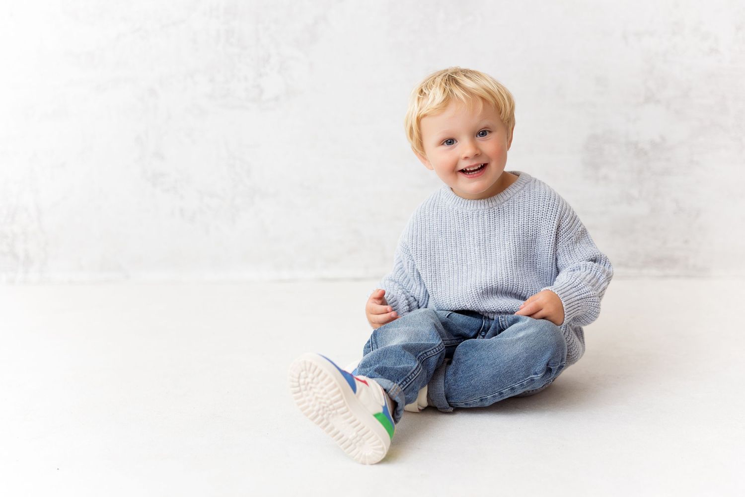 2 year old toddler wearing gray sweater and jeans sits laughing against white wall background. Classic Clean gap look.