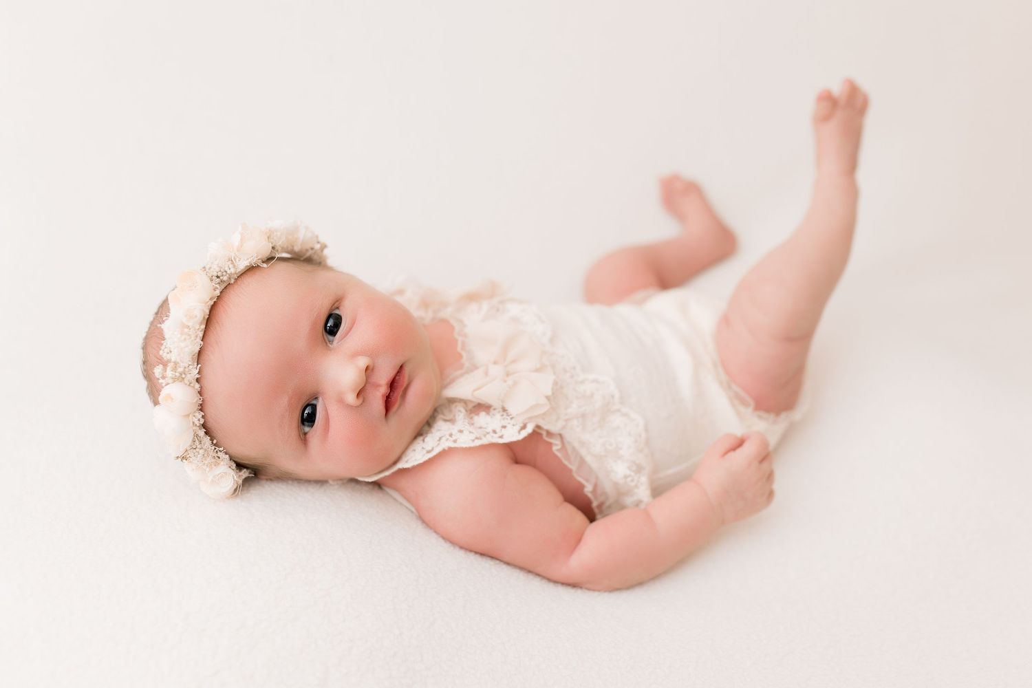 Baby in lace outfit lying on cream backdrop, awake newborn.