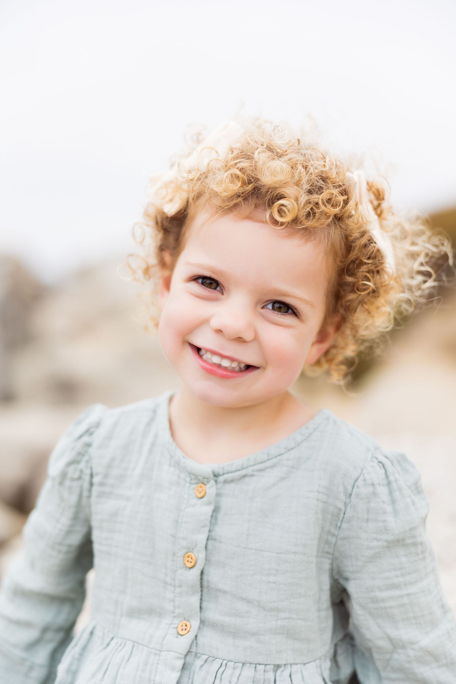 Young child wearing a light blue linen dress smiles brightly against a bright background.