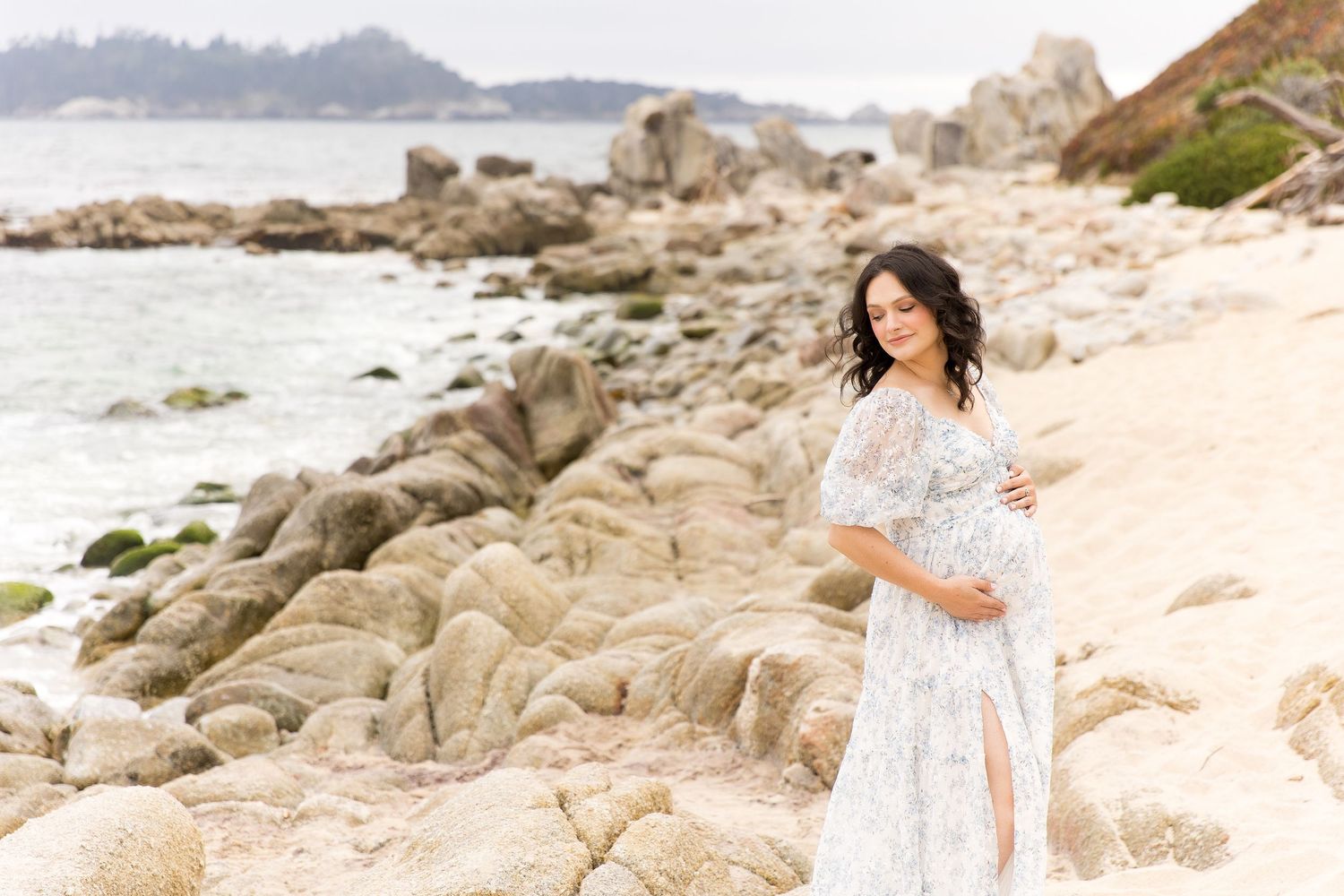 A pregnant mom in a flowy white dress walks barefoot along mossy rocks at a scenic beach during sunset.