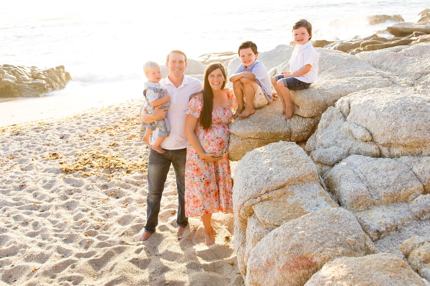 A family enjoys a sunset photoshoot together on a rocky beach with golden light filtering through.