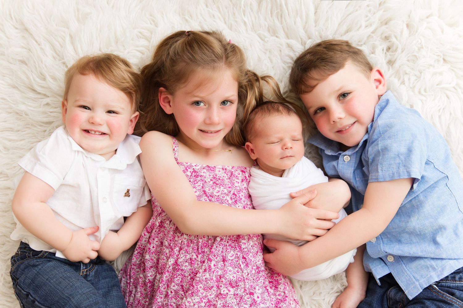Four children posed together on white fur rug with newborn baby sibling.